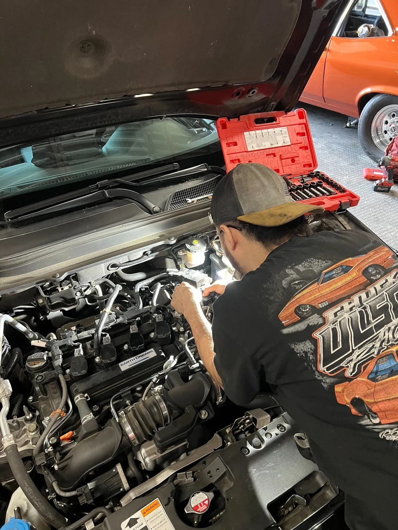 Person working on a car engine with tools in an open hood. A classic orange car is in the background. | Ashten's Total Auto Care