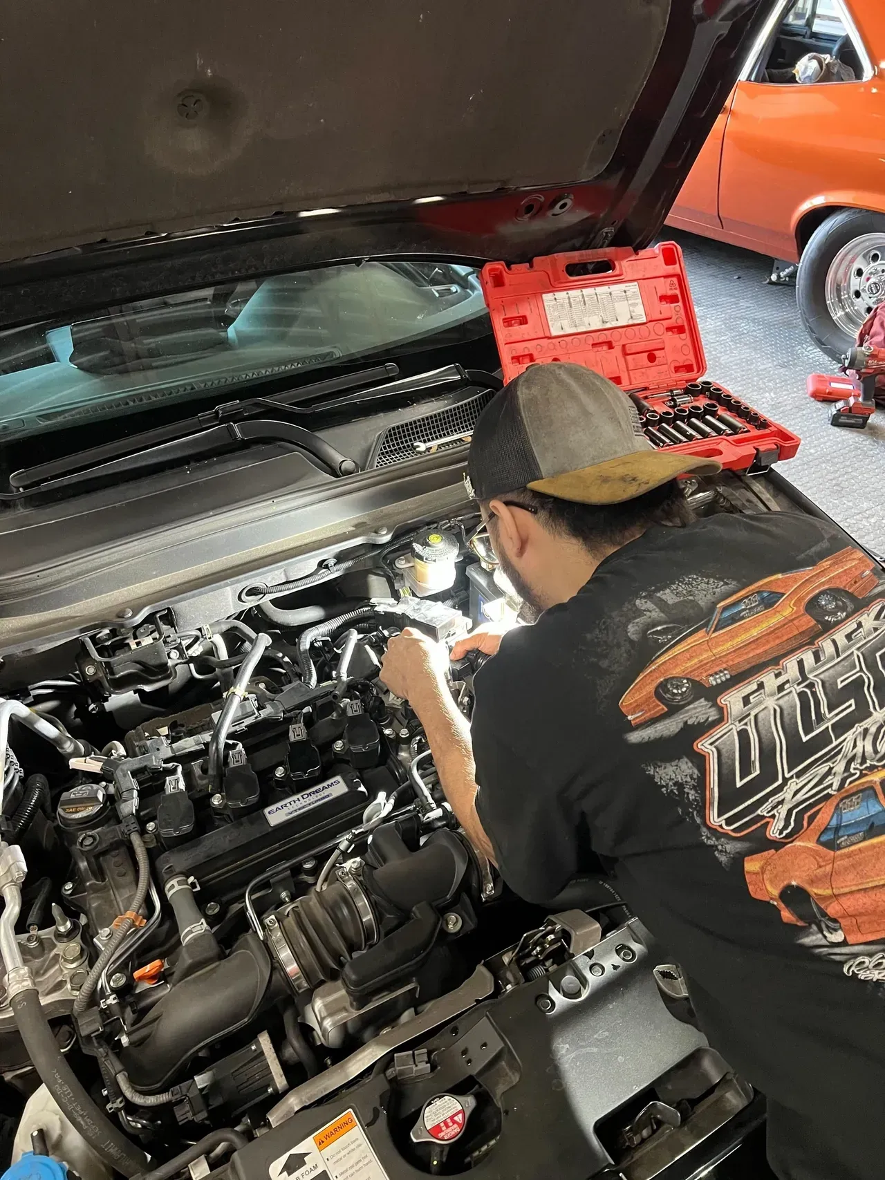 Person working on a car engine, under the open hood. A toolbox sits nearby in a garage. | Ashten's Total Auto Care
