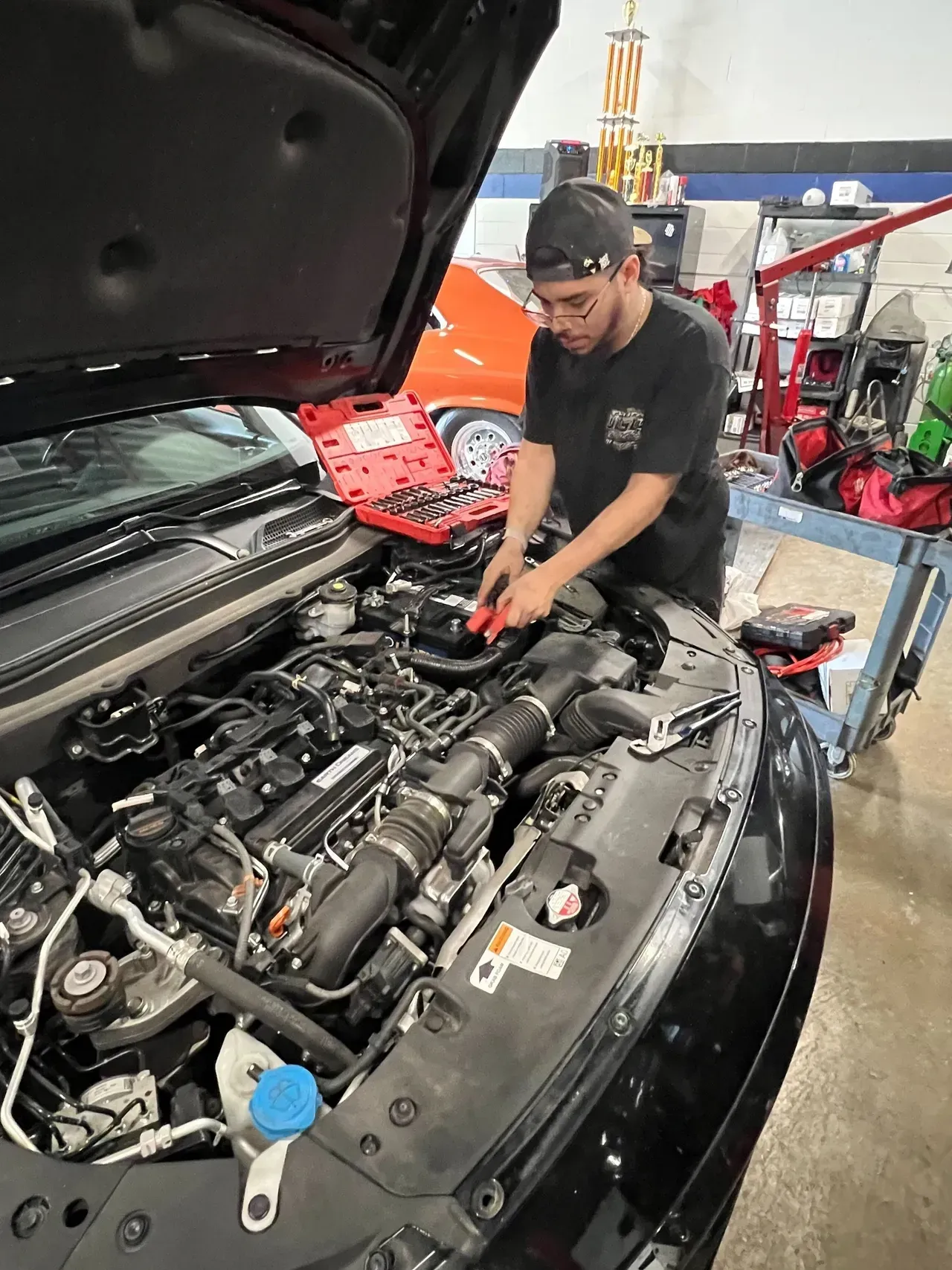 Mechanic working on car engine in a garage. Wearing a hat and dark shirt, tools nearby. | Ashten's Total Auto Care
