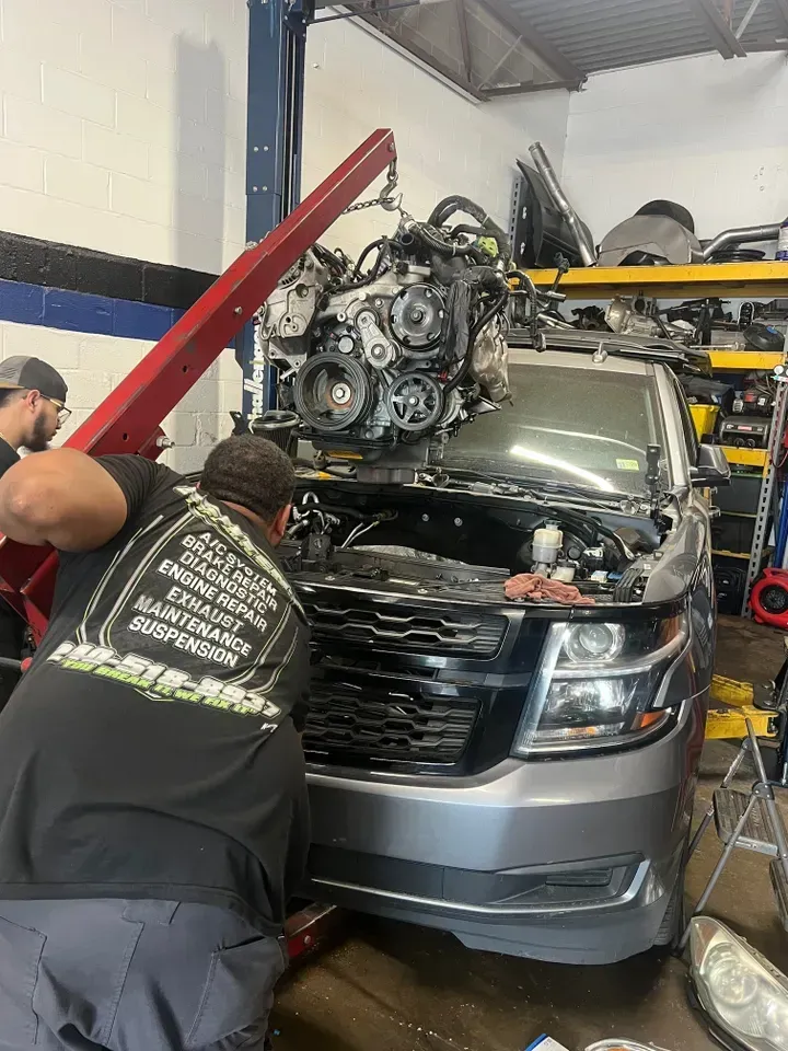 Mechanic removing engine from gray SUV in a shop. A crane holds the engine, another person assists. | Ashten's Total Auto Care