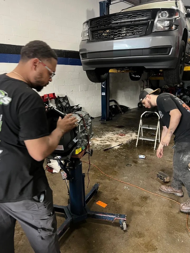 Two mechanics working on a car engine in a garage; a truck is raised above them. | Ashten's Total Auto Care