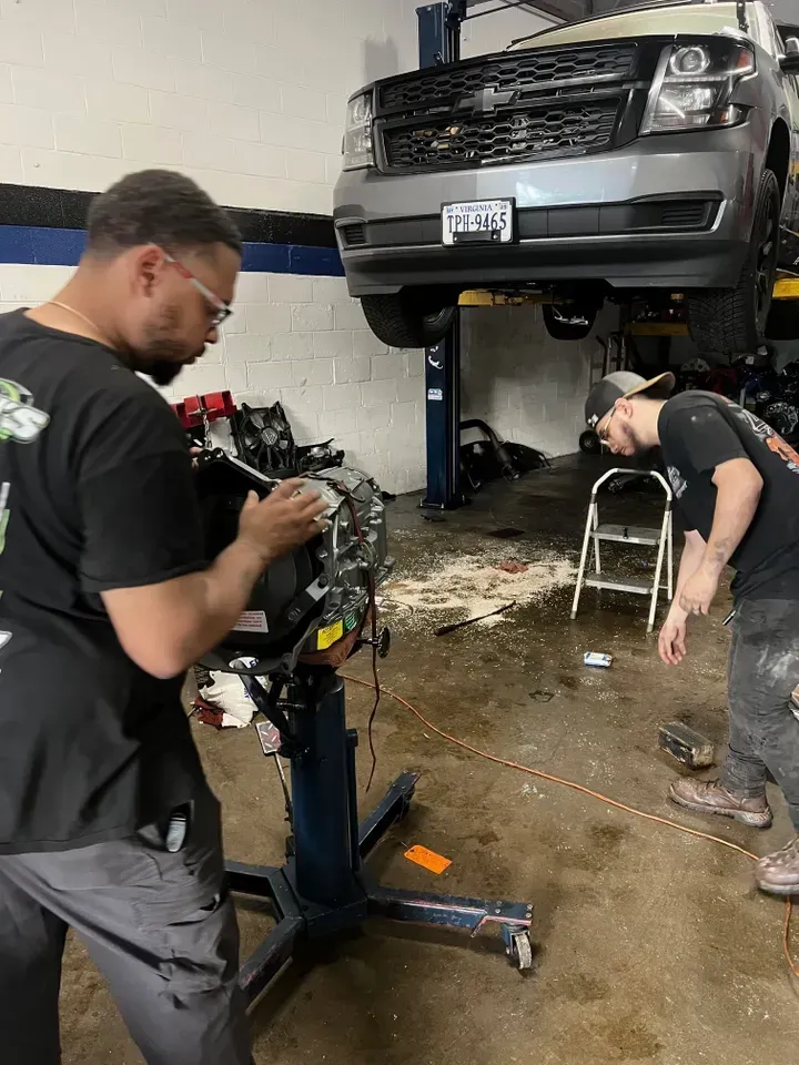 Two mechanics working on car parts in a garage, a car is on a lift. | Ashten's Total Auto Care