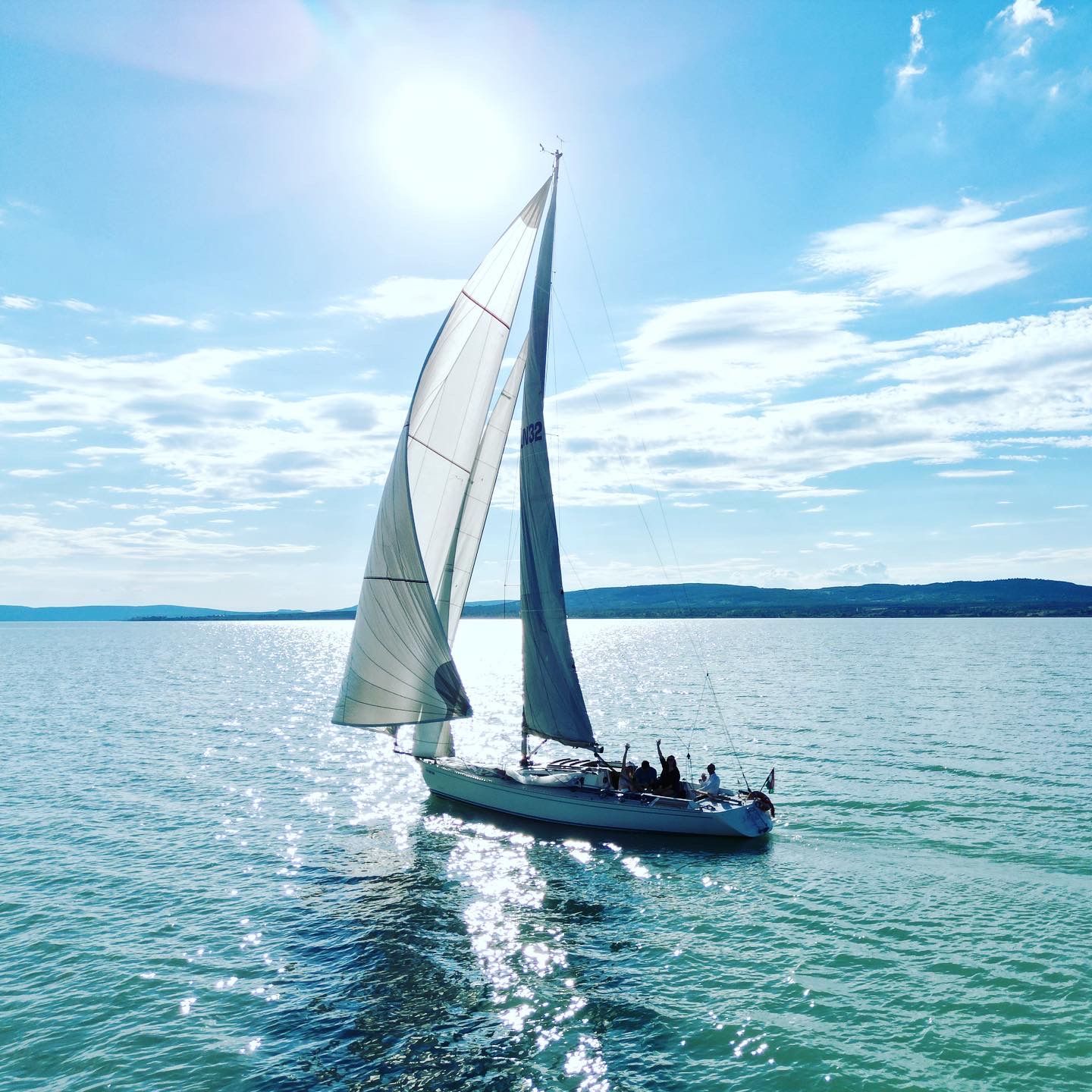 Sailboat on calm water, bright sun overhead. People on deck. Blue water and sky, distant shoreline.