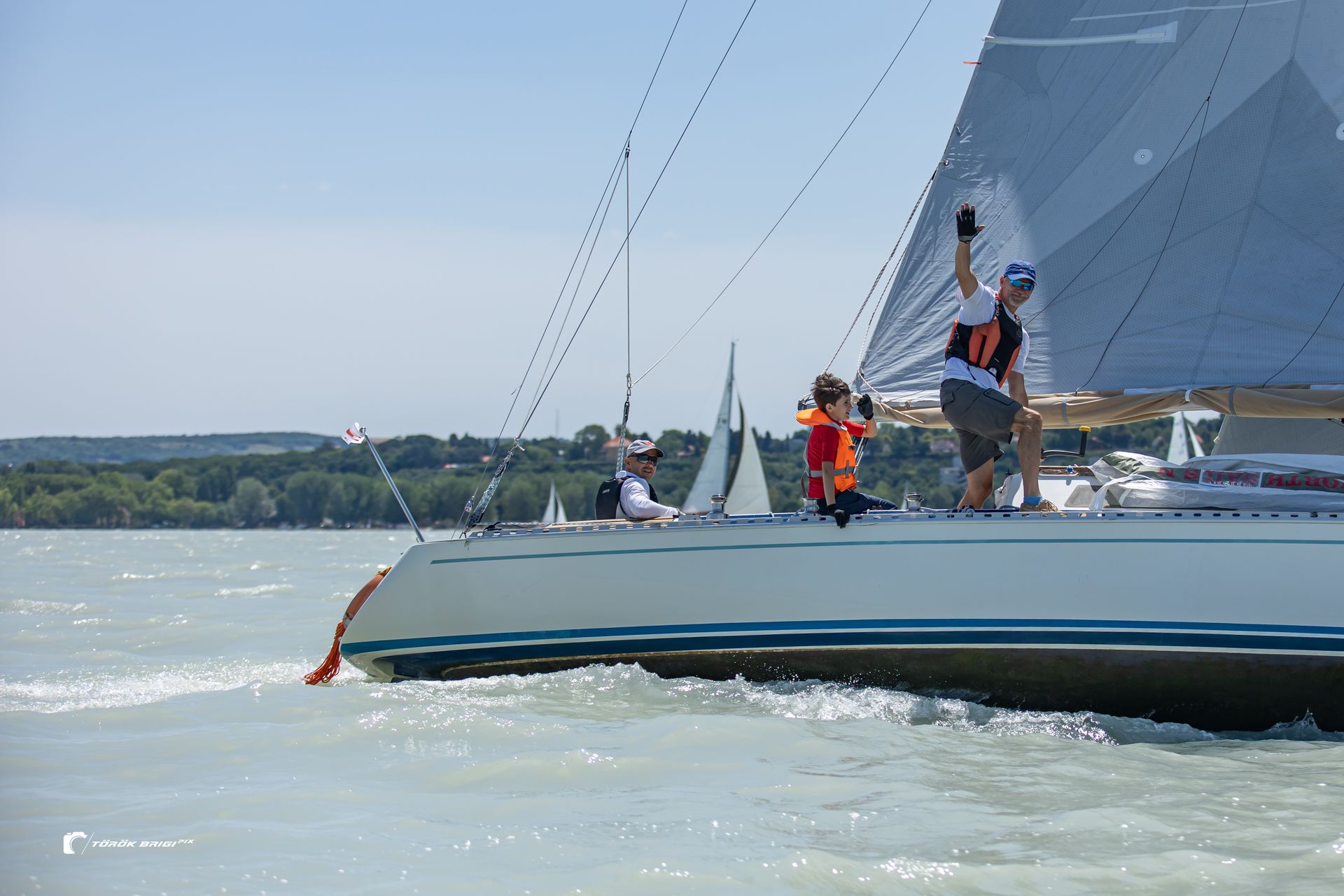 Sailboat on a sunny day with people on deck waving. Blue water, blue and white boat, green shoreline.