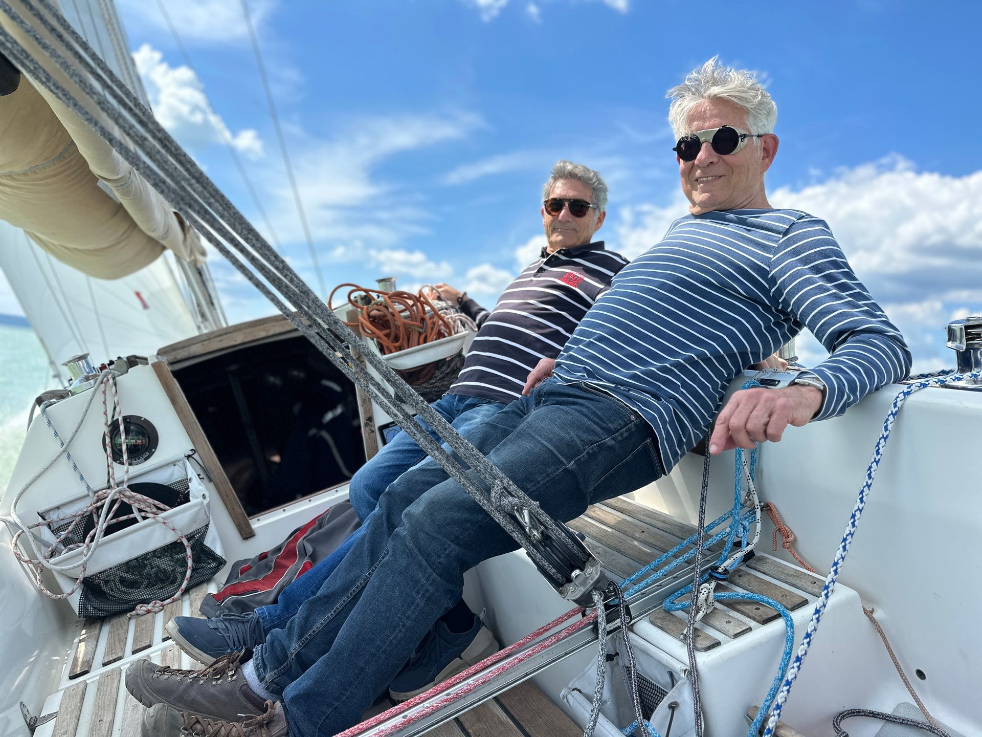 Two men on a sailboat, leaning back. Sunny day with blue sky. One in striped shirt, one in dotted.