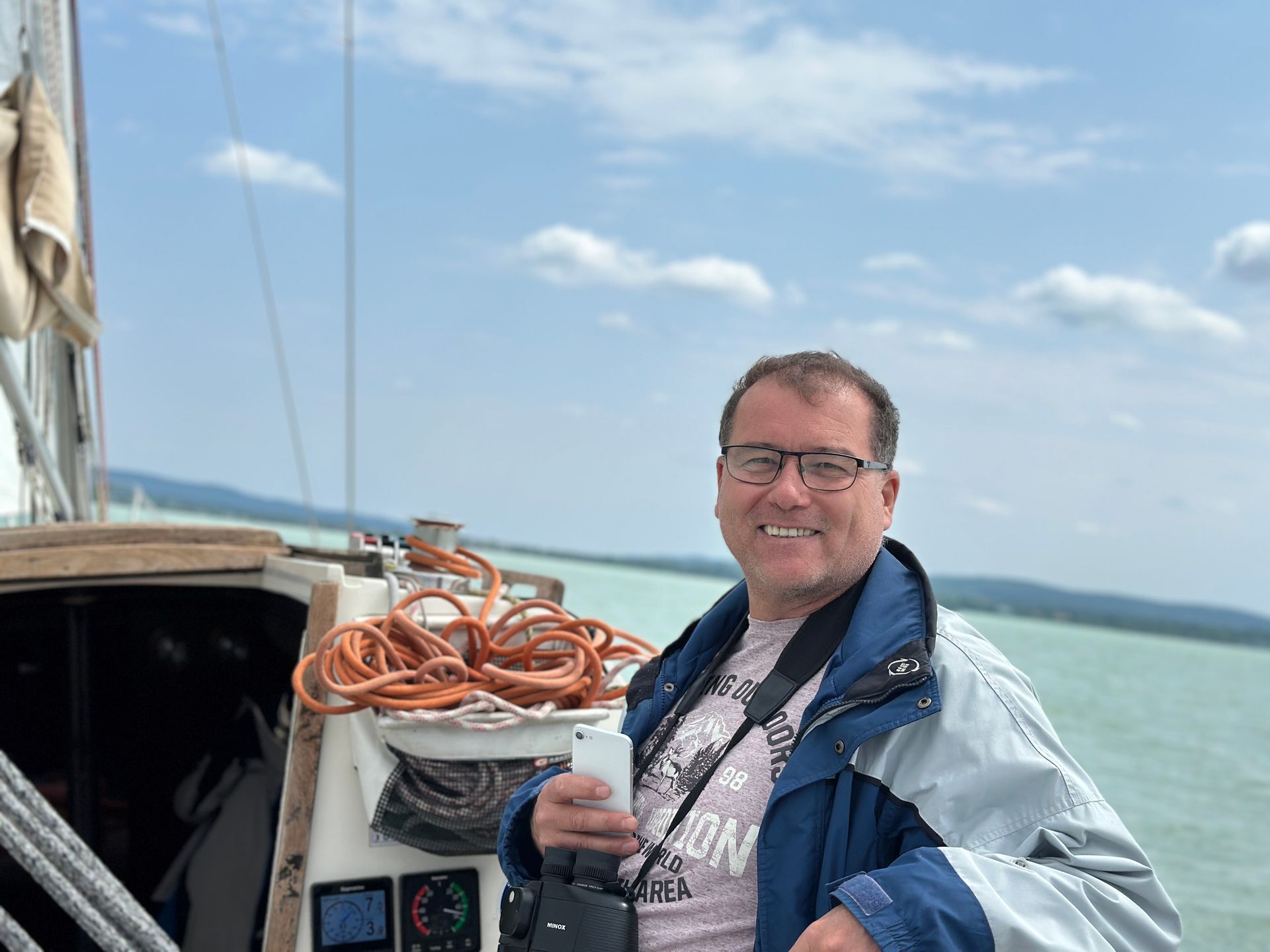 Man on a sailboat smiling, holding a phone. Blue jacket, sea background.