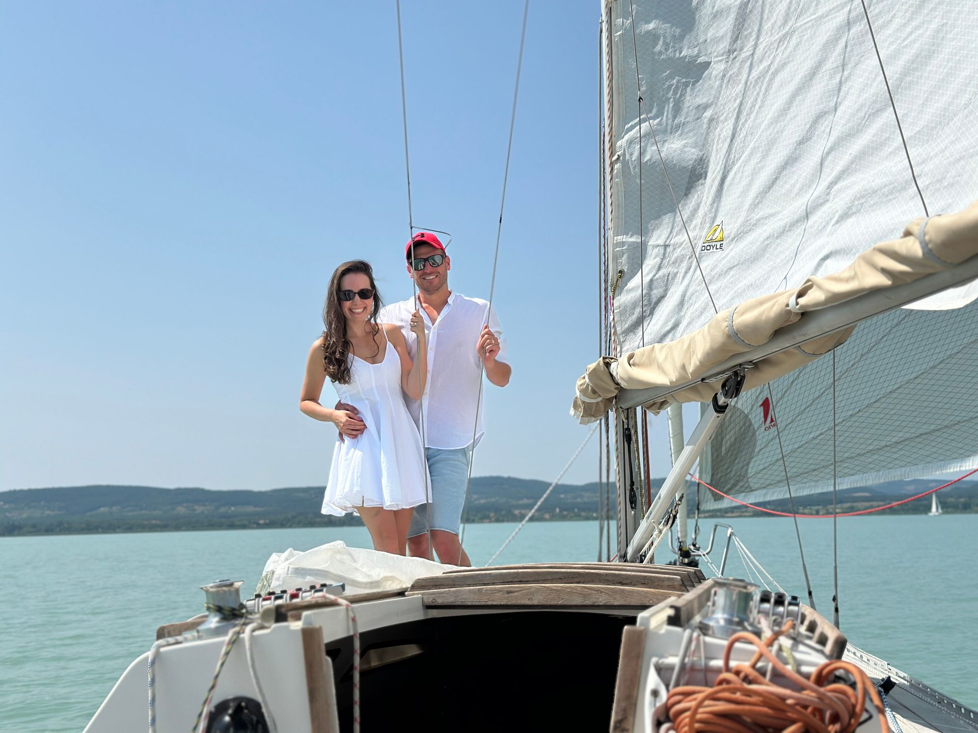 Couple on sailboat; woman in white dress, man in cap, smiling on sunny water.