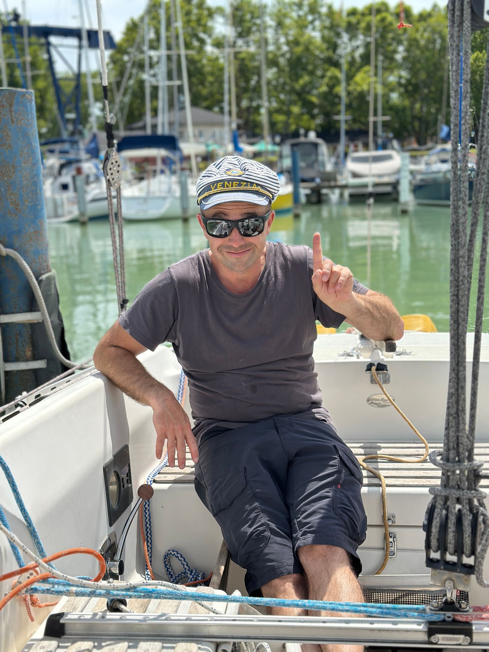 Man on sailboat wearing captain's hat points up. He sits in cockpit, marina in background.