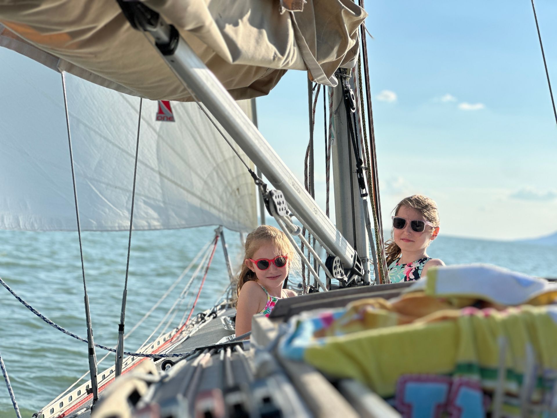 Two girls in sunglasses on a sailboat; blue water and sky, smiling, looking at the camera.