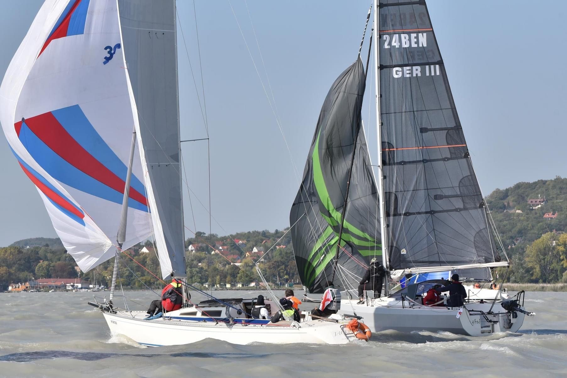 Two sailboats racing on a lake. One has a colorful spinnaker, the other a black one. Crew members are visible.