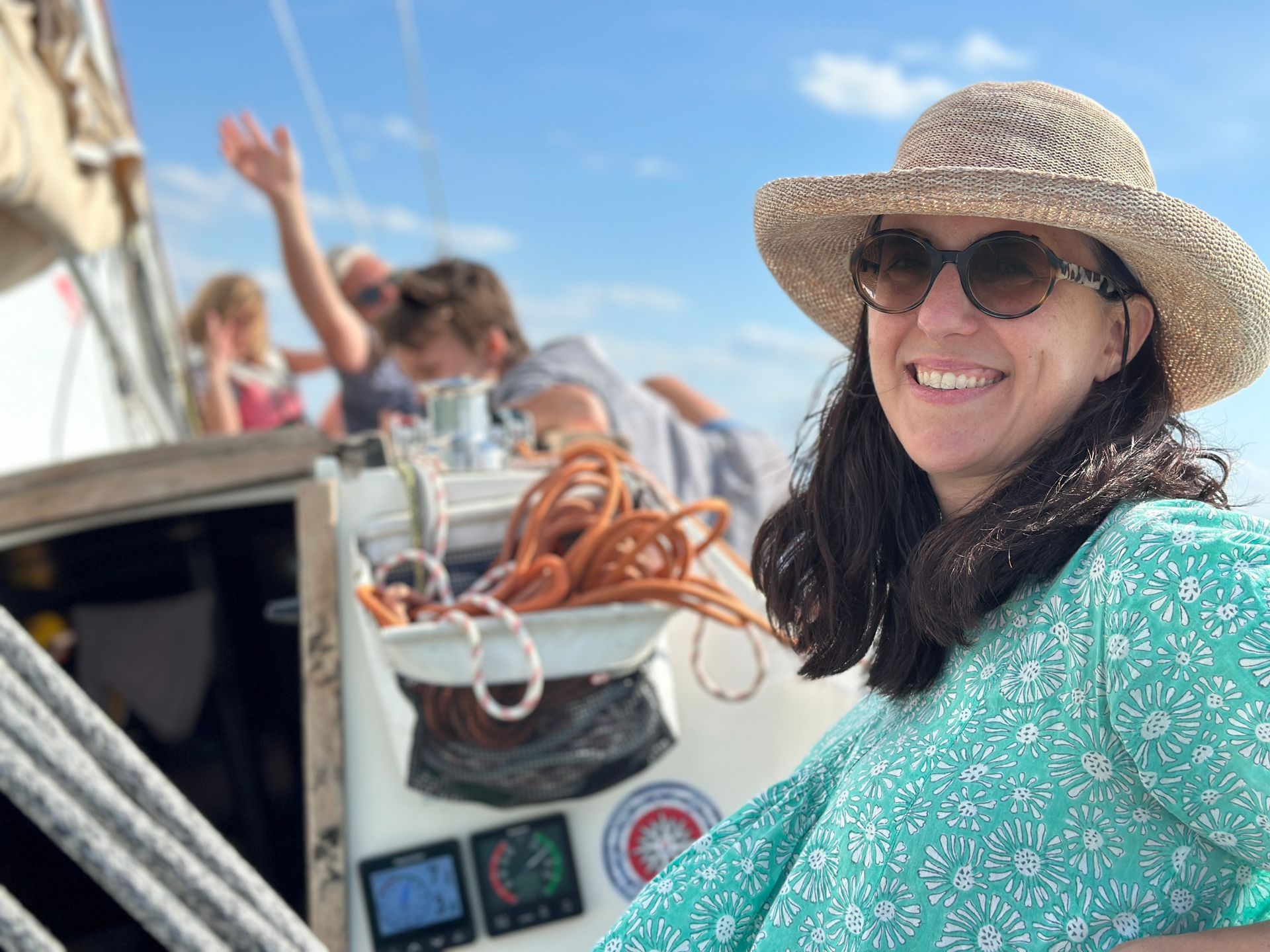 Woman in a straw hat smiles on a sailboat with others in the background, sunny day.