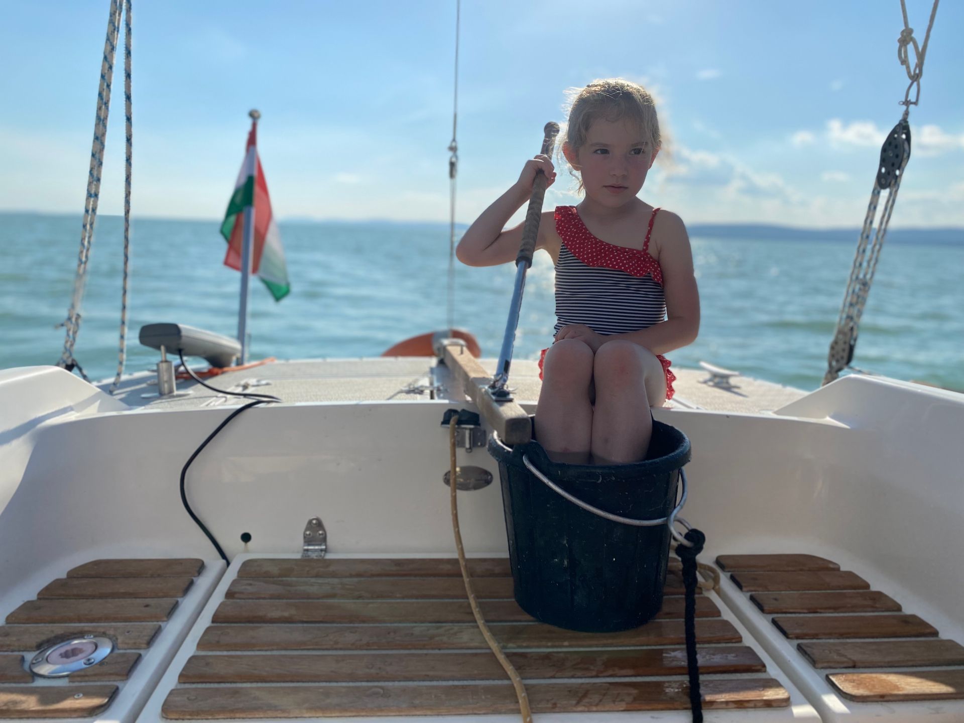 Girl in swimsuit sits in a bucket on a sailboat, holding a pole. Hungarian flag waves in the background.