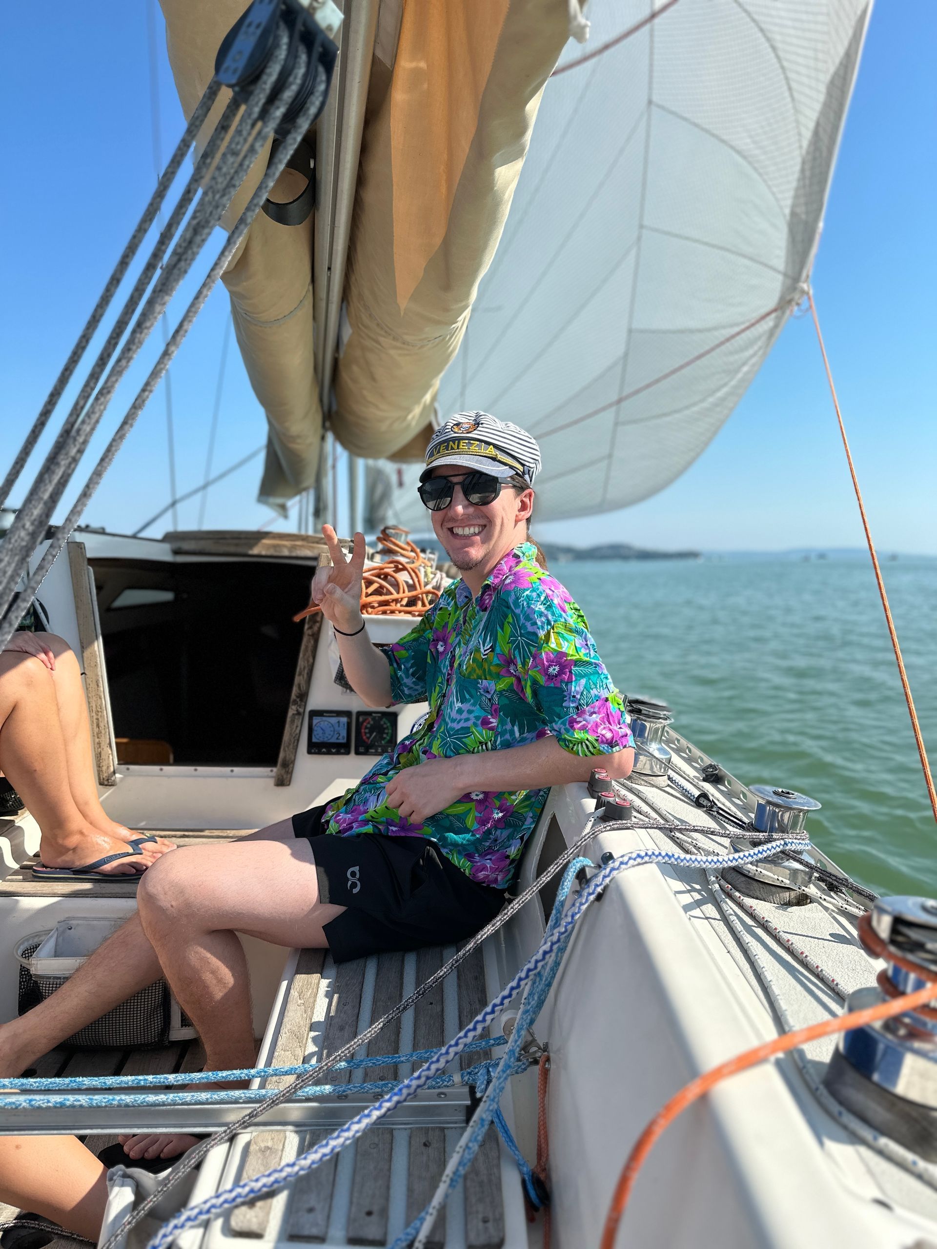 Man in floral shirt and striped hat waves from a sailboat on sunny water.