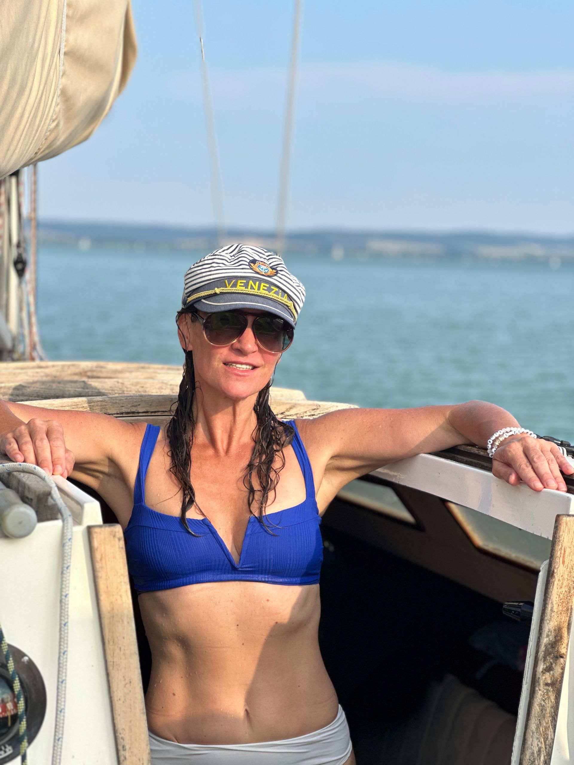 Woman on sailboat wearing a captain's hat and blue bikini, arms resting on boat edge.