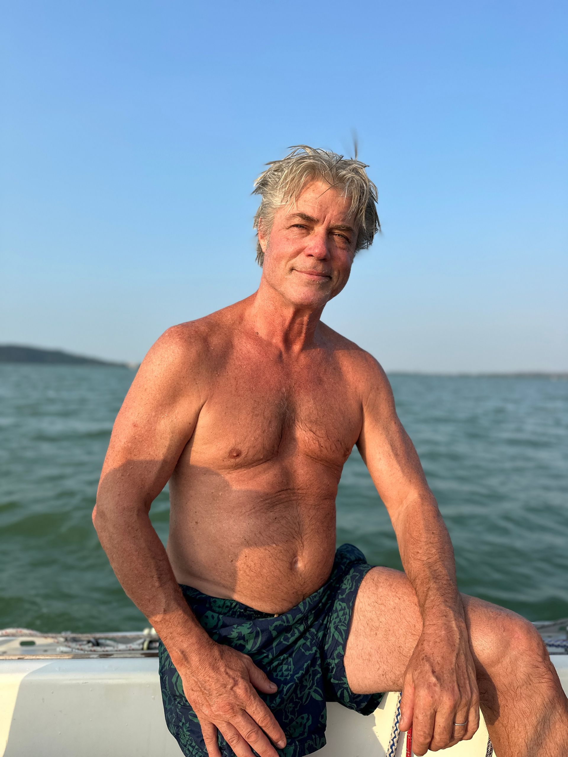 Man, shirtless, on boat in water. Blue sky, dark blue swim trunks.