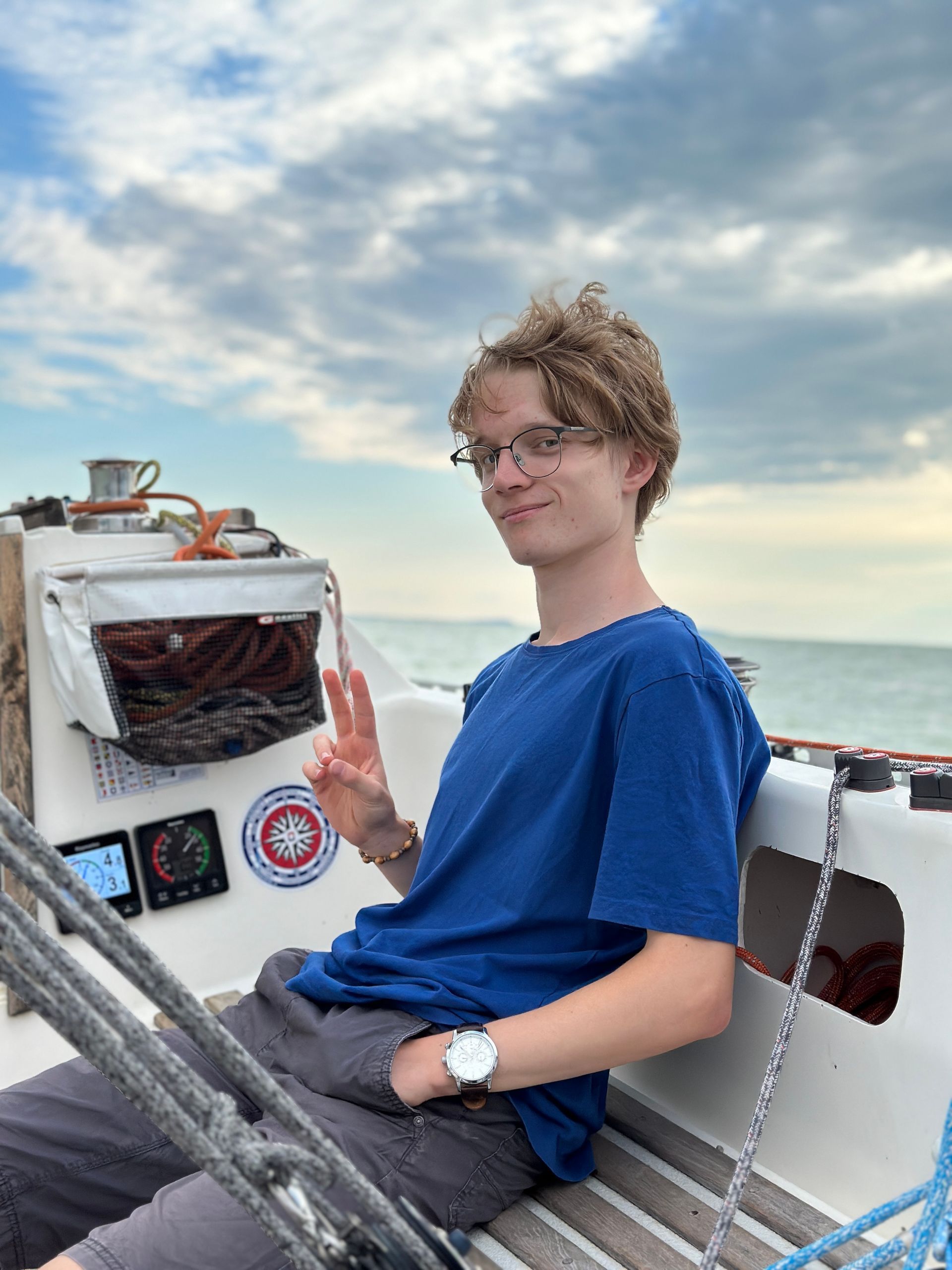 Young man with glasses on a sailboat, holding up peace sign, smiling, sea in background.