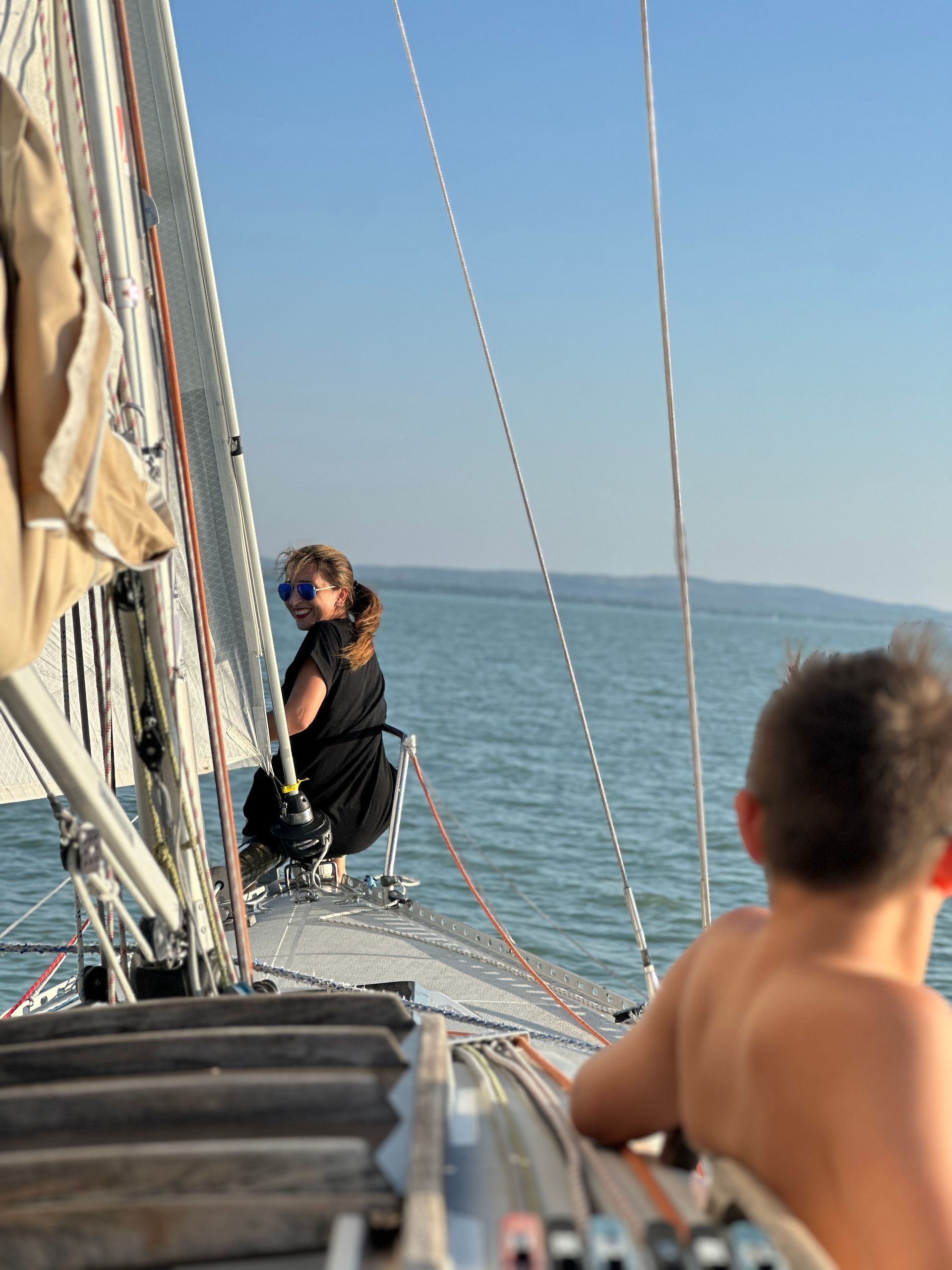 Woman on sailboat looks back, boy in foreground; blue sky, water.