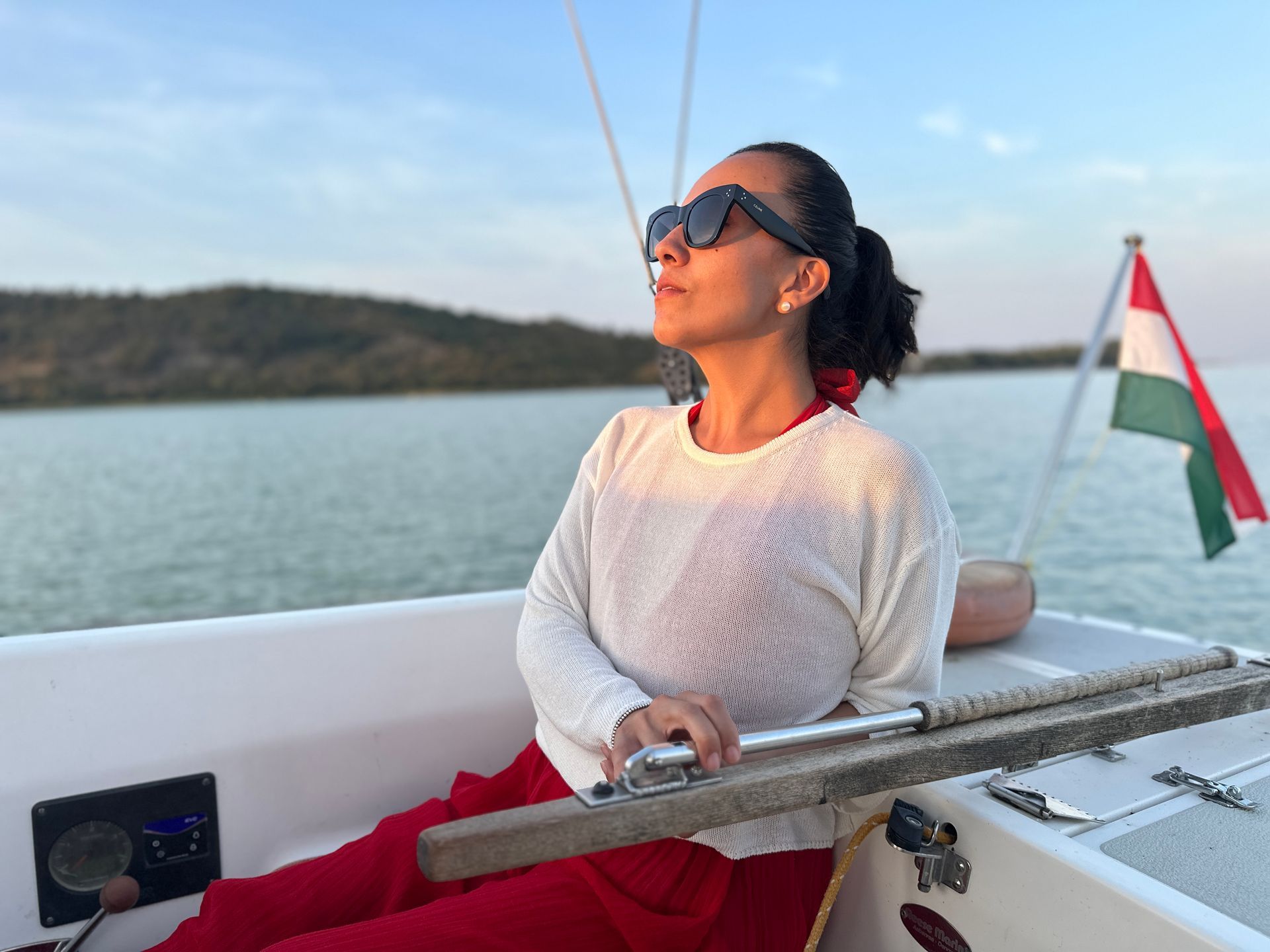 Woman sailing on a boat, looking up, wearing sunglasses and red pants. Hungary flag in the background.