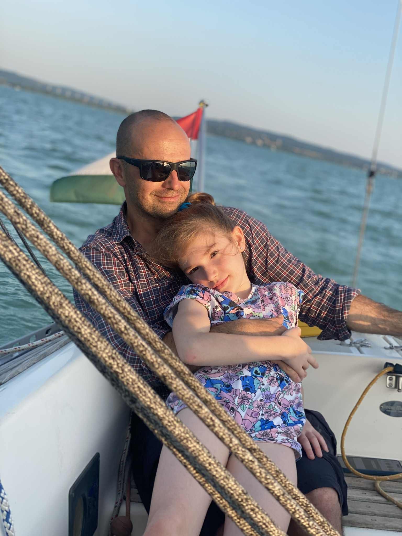 Man and girl on a sailboat, embracing, looking at the camera. The sea and land in the background.