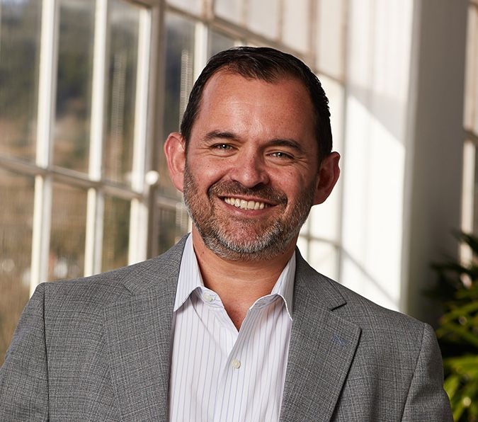 Man in gray blazer smiles in a bright indoor setting.