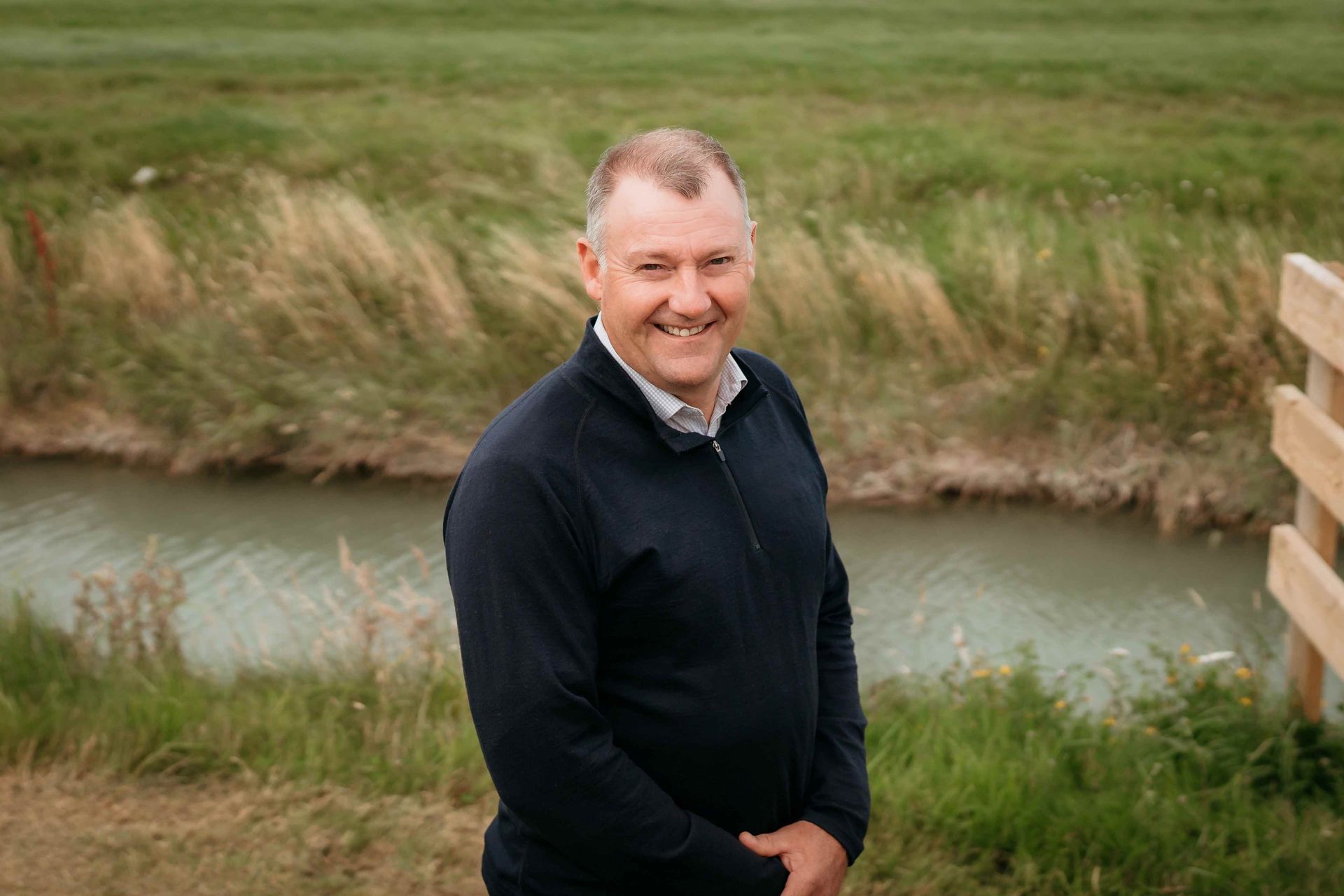 Man in a DairyNZ shirt smiles in a green field with black and white cows grazing in the background.