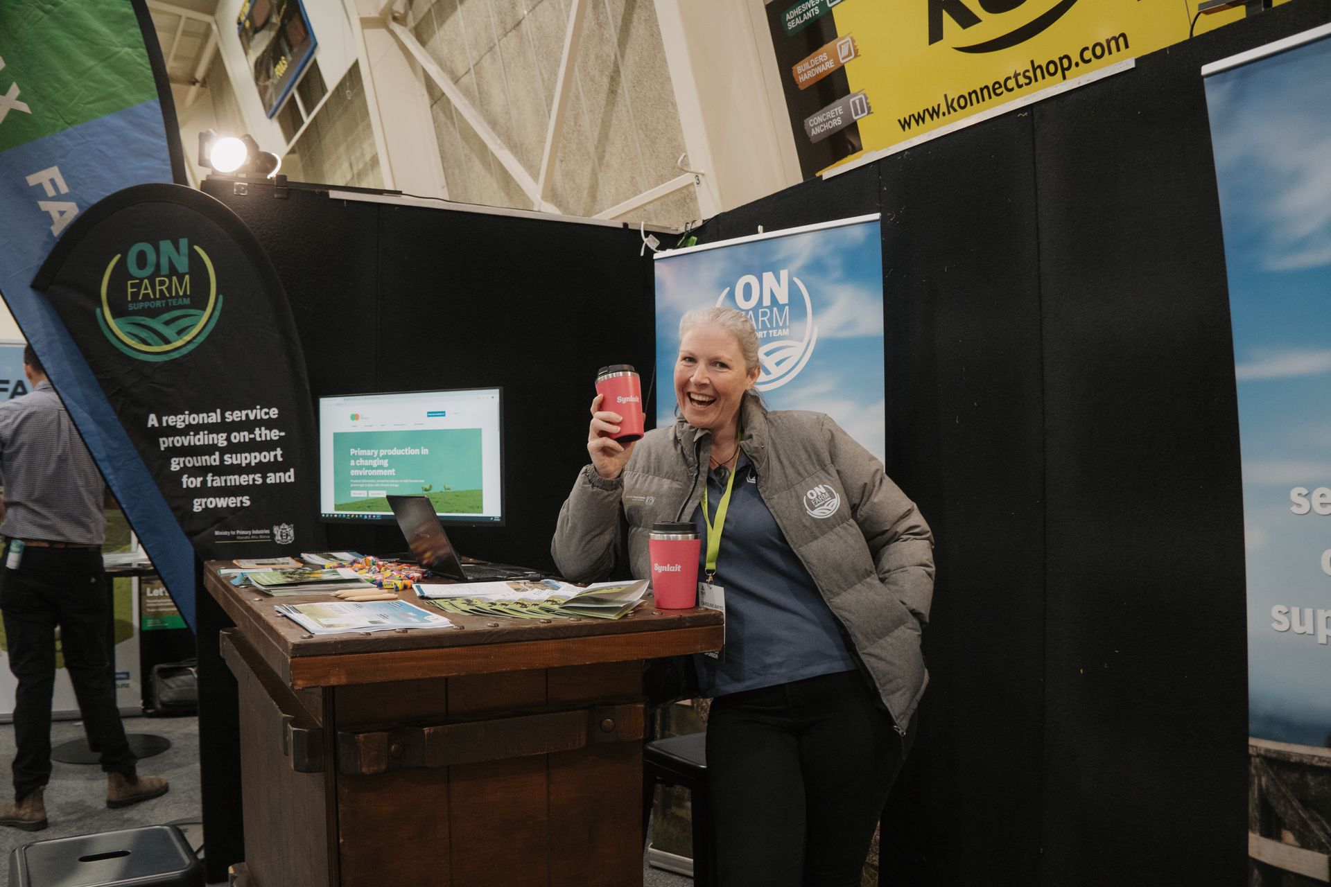 Woman smiling at a trade show booth, holding a red drink can, wearing a puffy jacket.