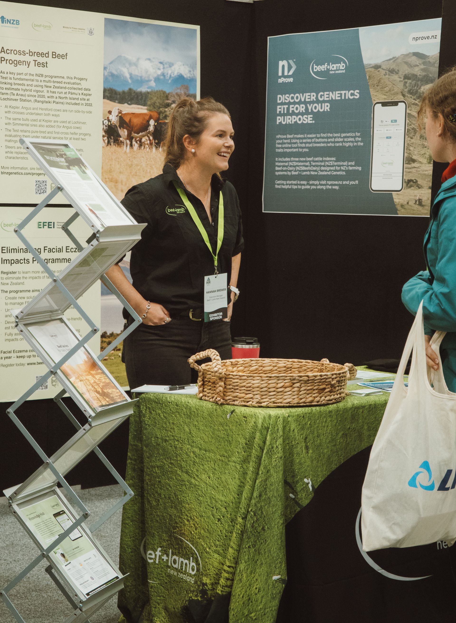 Woman at a trade show booth, talking to another person, green tablecloth, brochures, and posters.