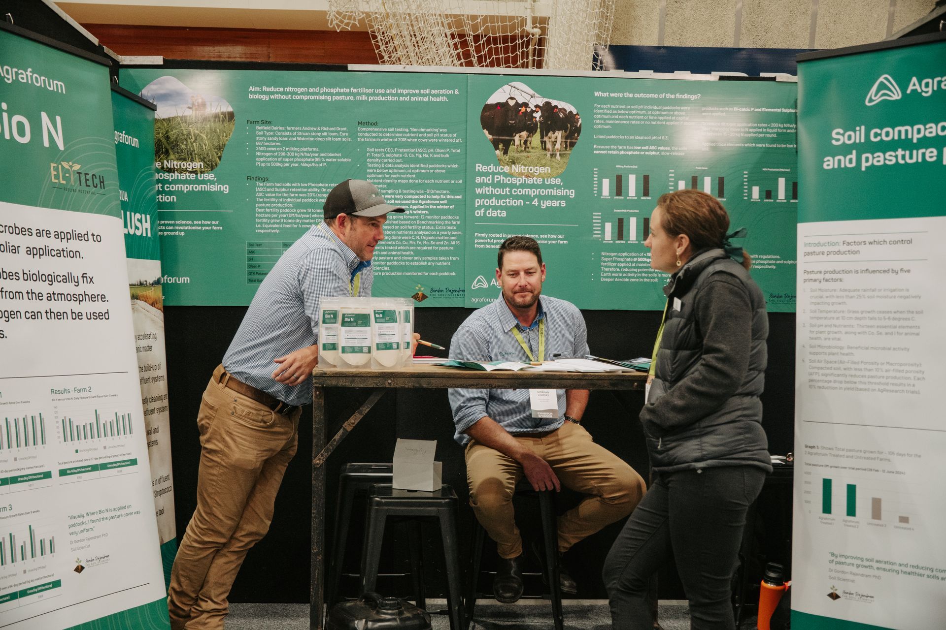 Three people at a trade show booth, discussing materials. Green backdrop with charts and text.