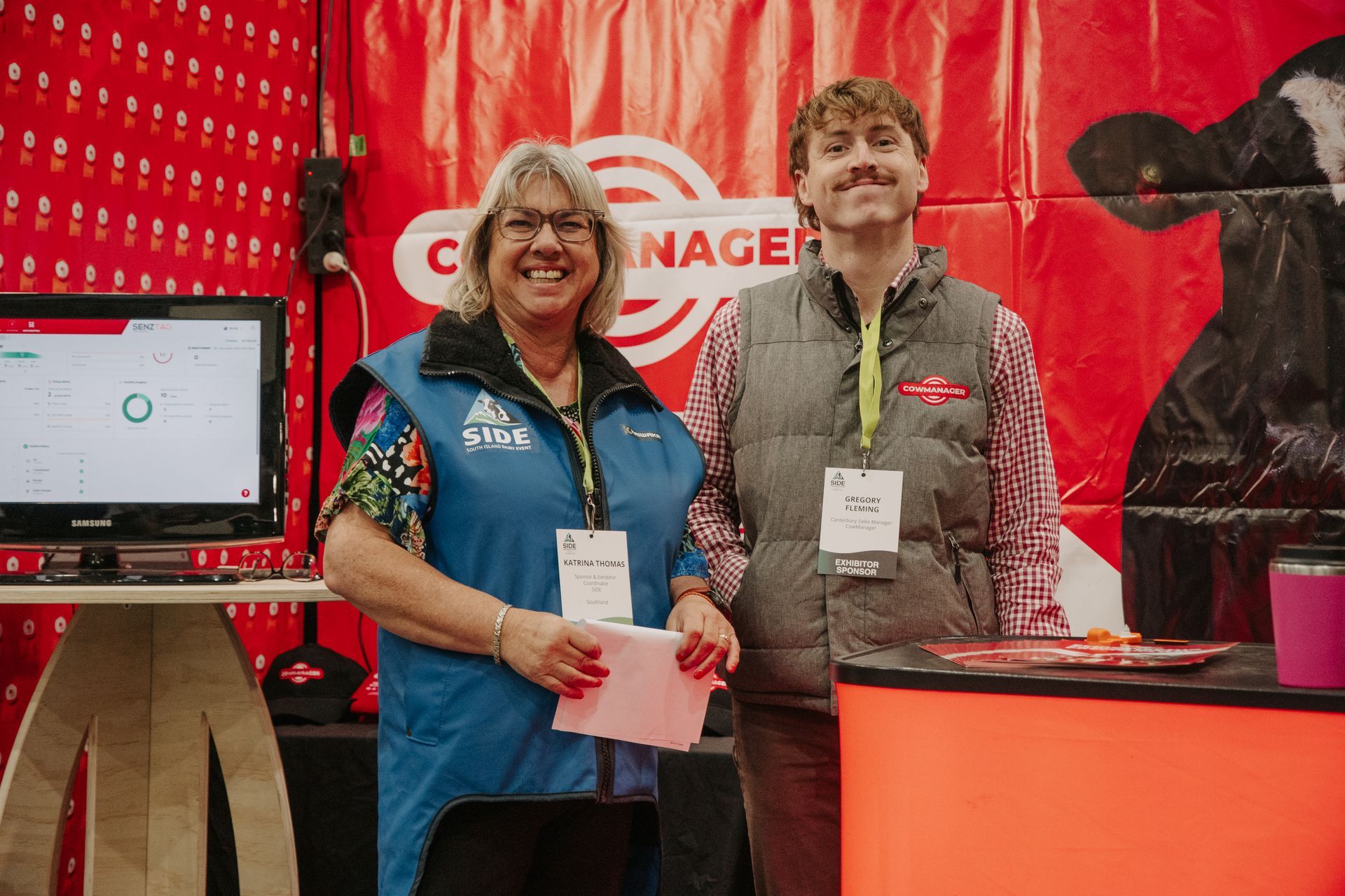 Two people at a booth with a screen. Woman in blue vest smiles, holding papers. Man in grey vest smiles. Red background.