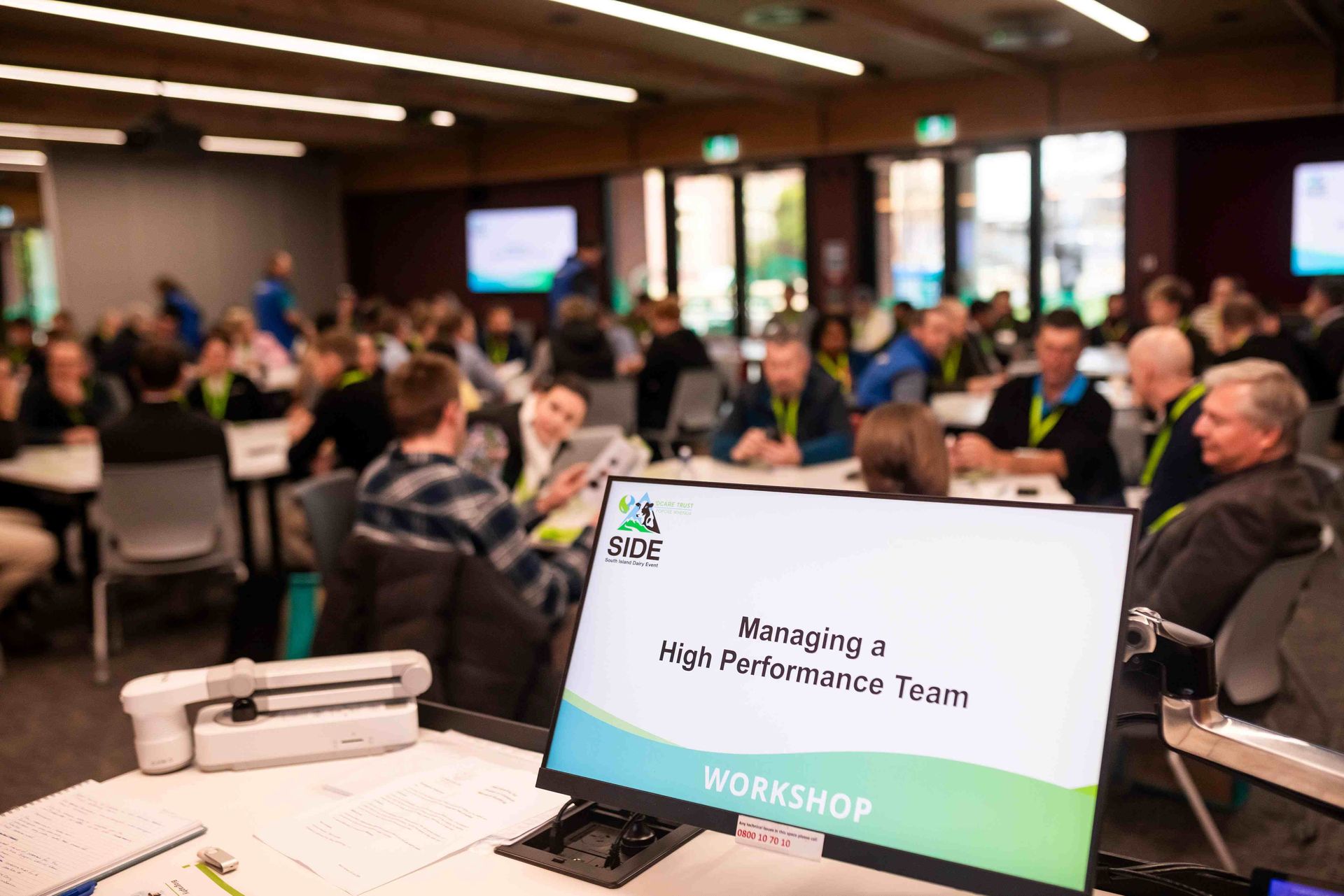 Workshop in a large room, people at tables. A screen in the foreground says 