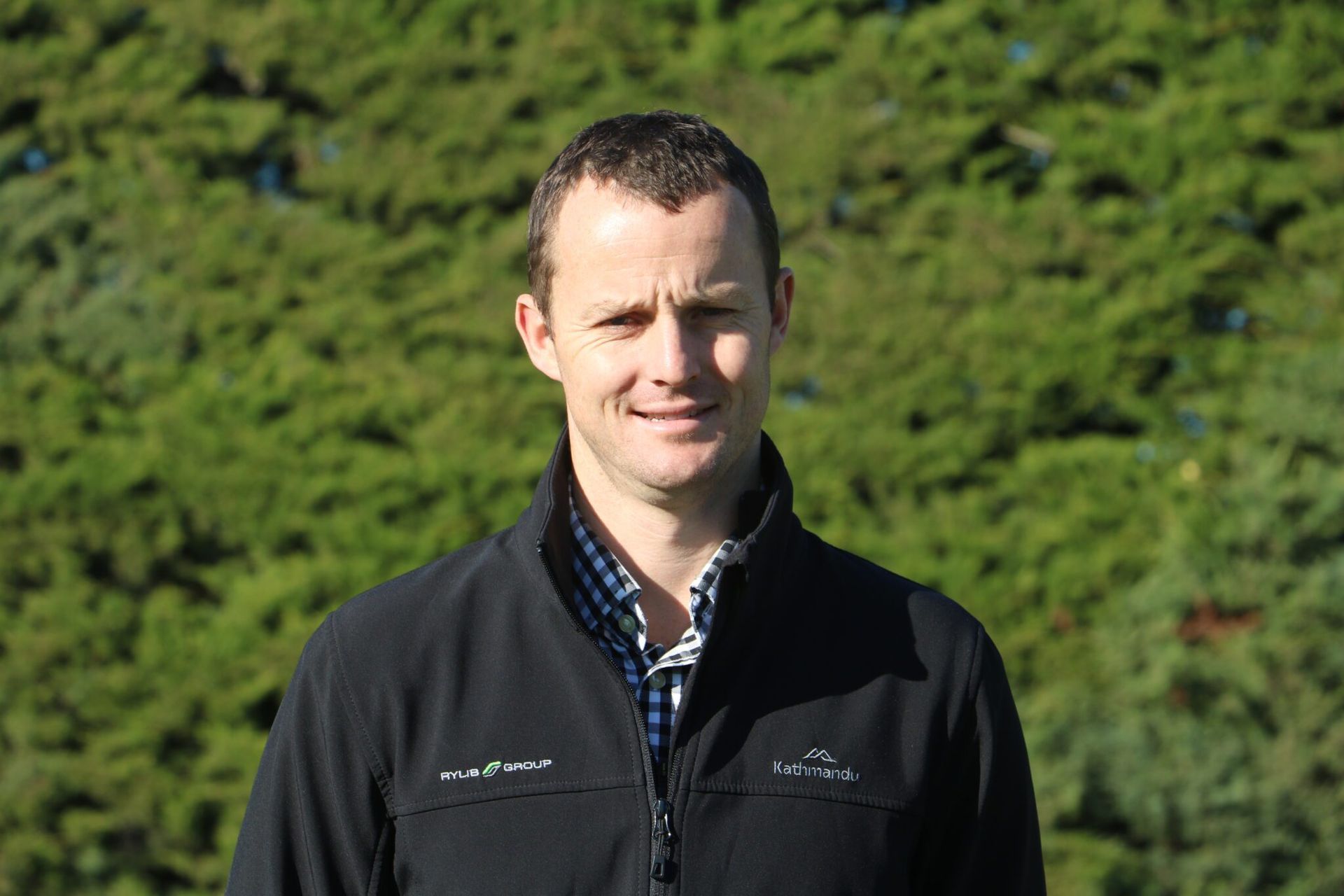 Man in black jacket smiling outdoors in front of green foliage.