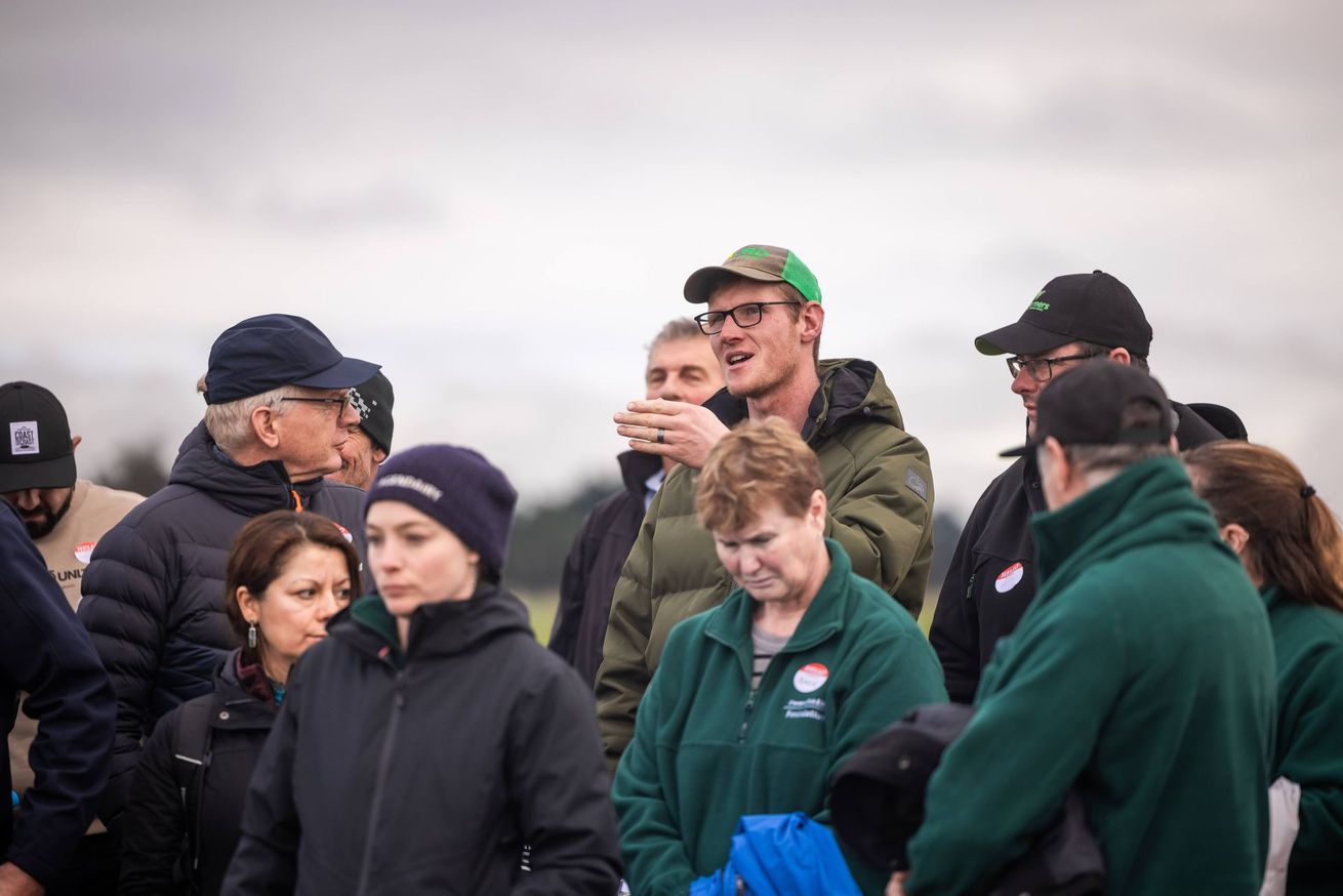 Group of people outdoors, some looking at a man speaking, cloudy sky.
