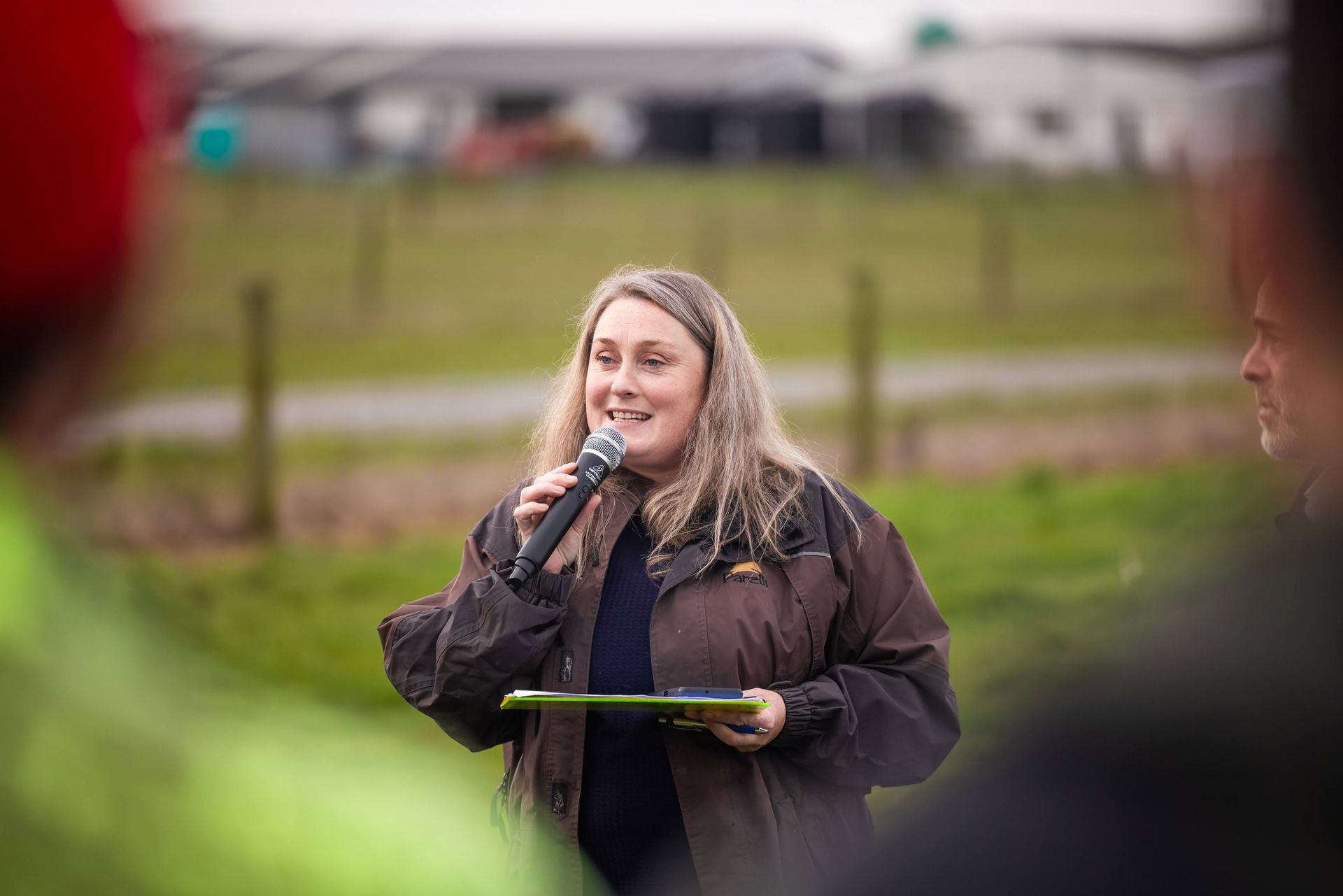 Woman speaking into a microphone outdoors, holding a clipboard, brown jacket, people in the blurred foreground.