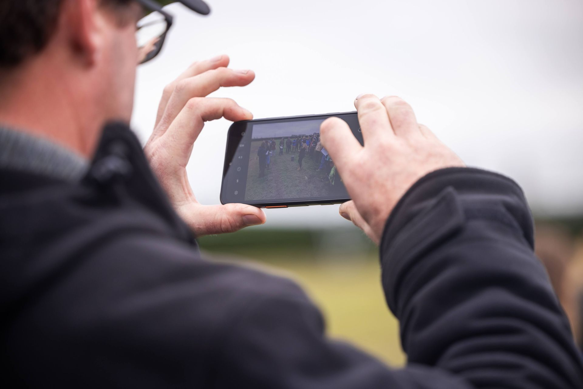 Person holding a smartphone, taking a photo outside, hands in frame, blurry background.