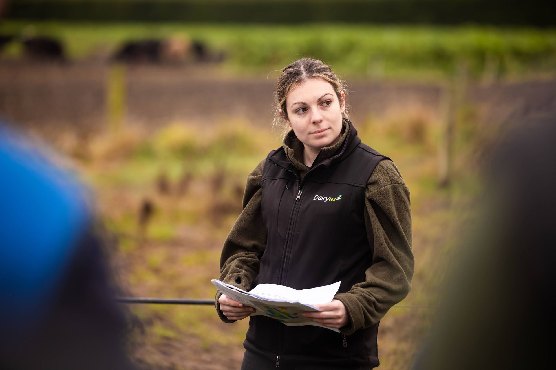 Woman in vest, holding papers, giving a presentation outdoors. Field and foliage in background.
