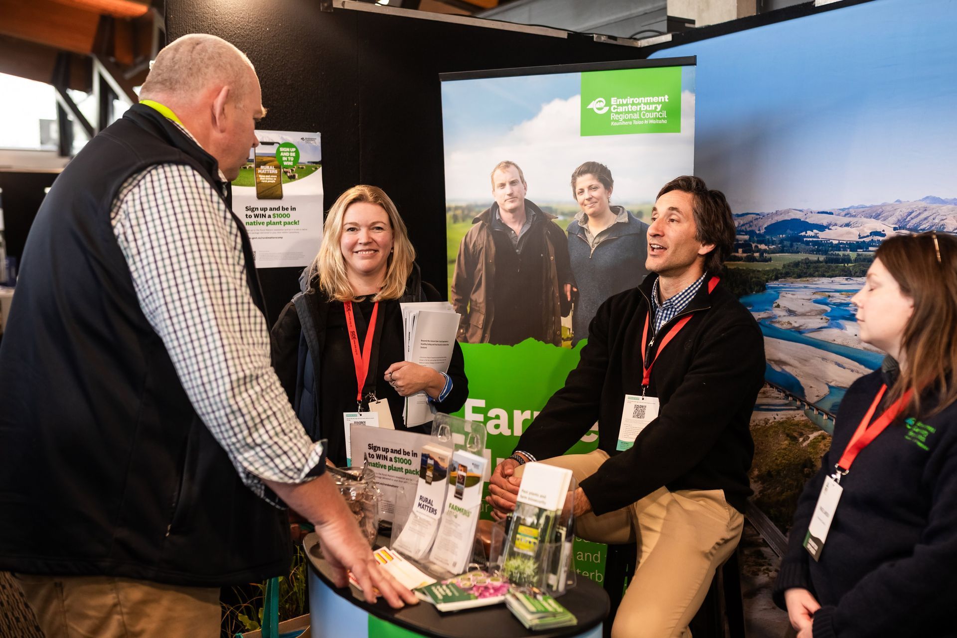 People at a booth, discussing products. Display of products on a table. Posters in the background.