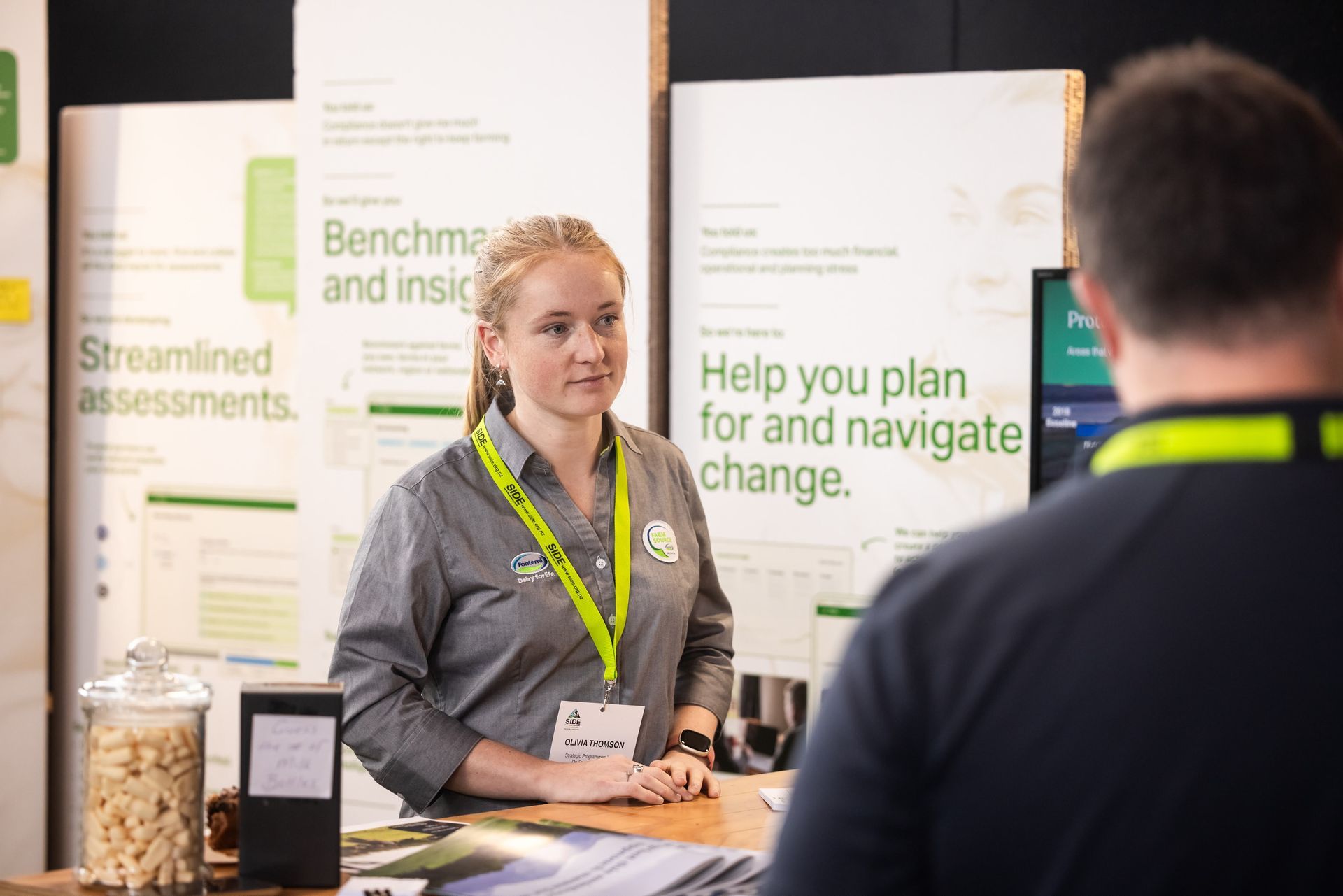 Woman in gray shirt at a booth, talking to a man. Signage with green and white text in the background.