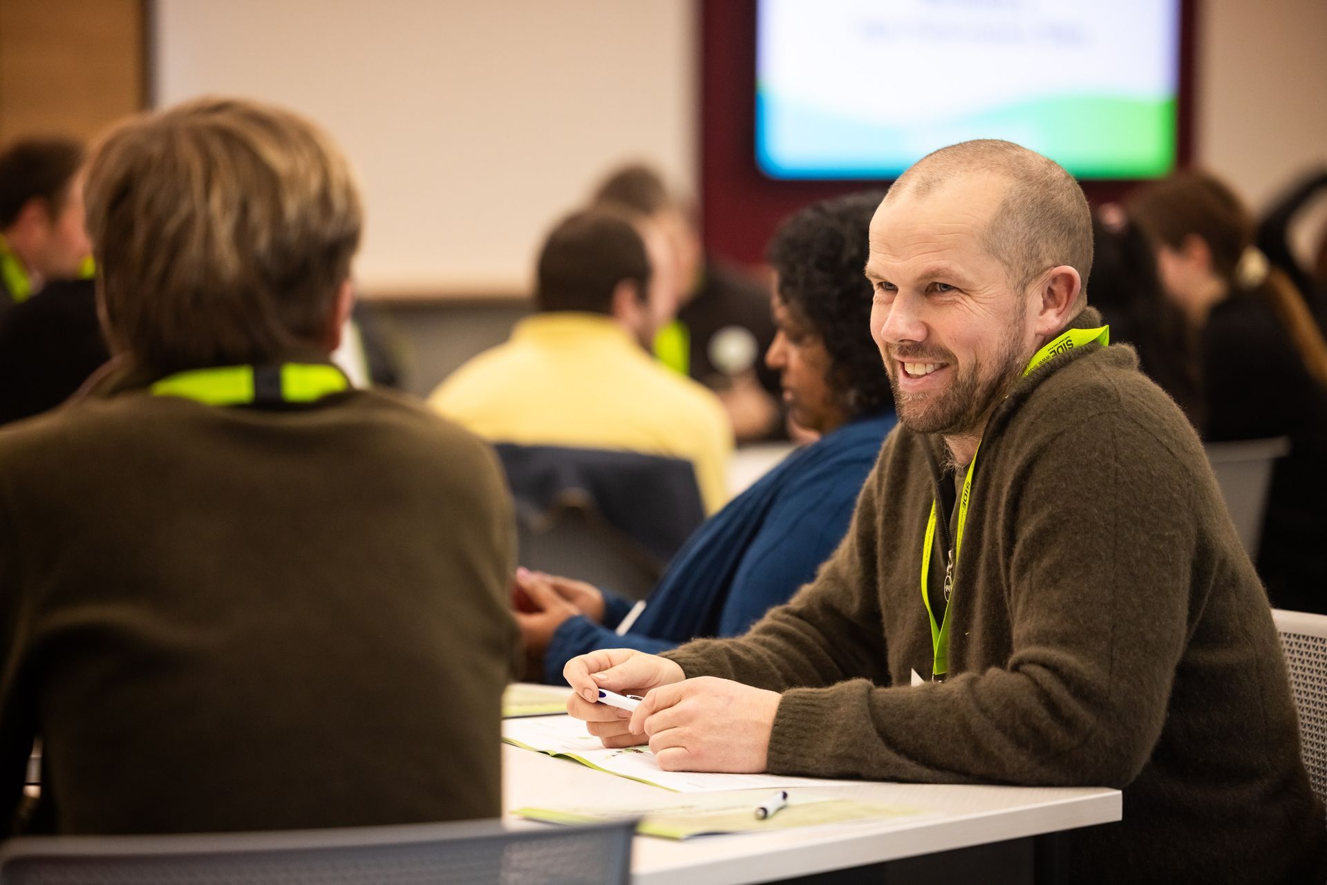 Man smiles, talking at table with others in meeting.
