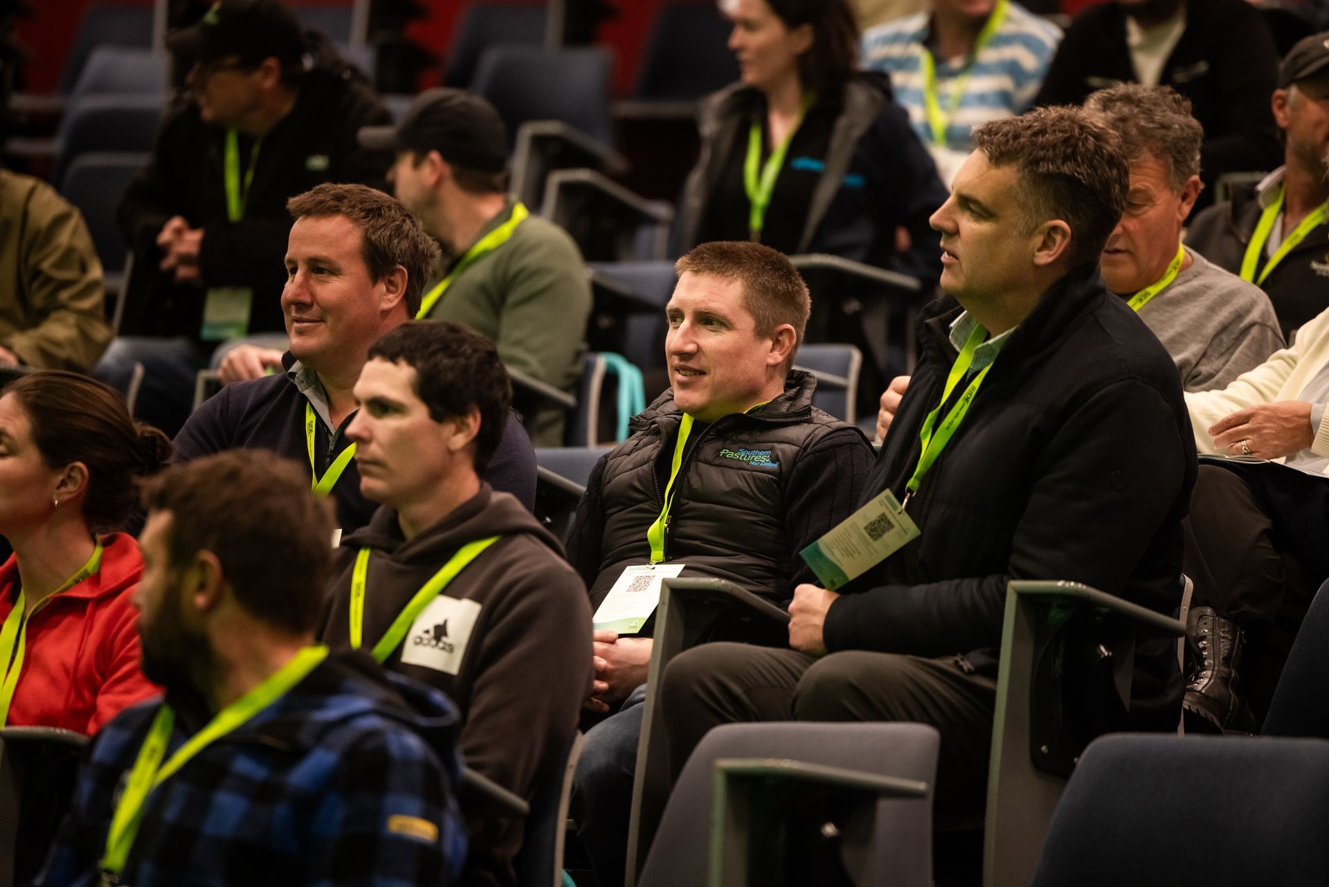 Audience members in an auditorium, listening attentively. Some wear lanyards, and jackets.