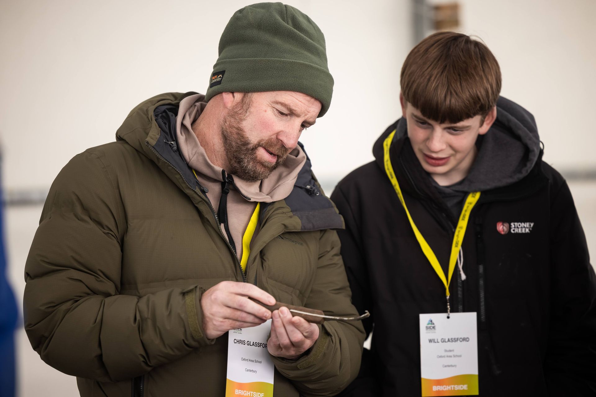 Man in green hat and boy examine small object, both wearing lanyards, indoors.