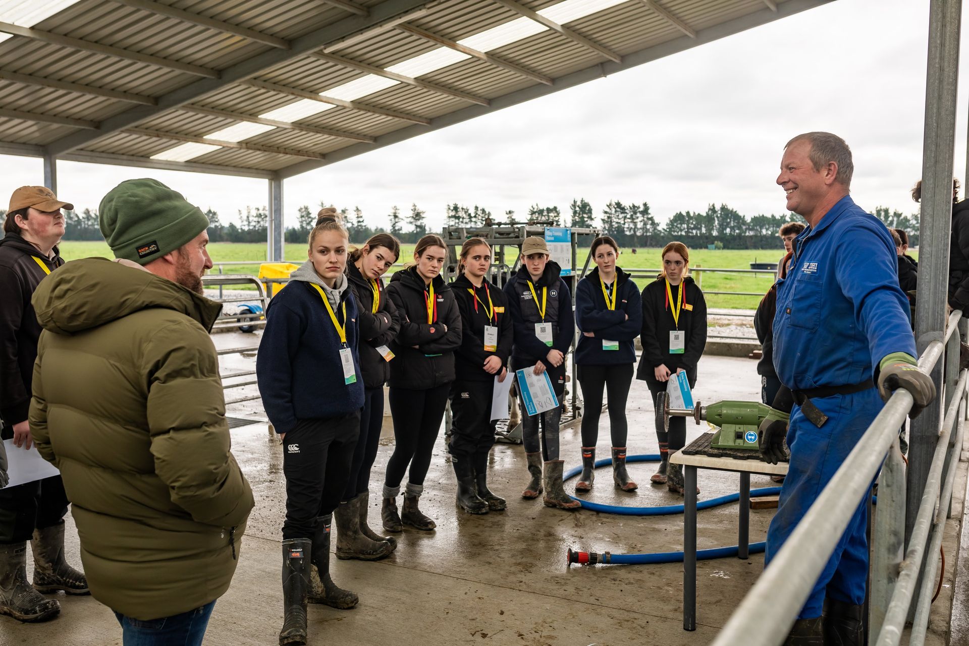 Group of people listening to a man in blue overalls outdoors under a covered area, surrounded by equipment.