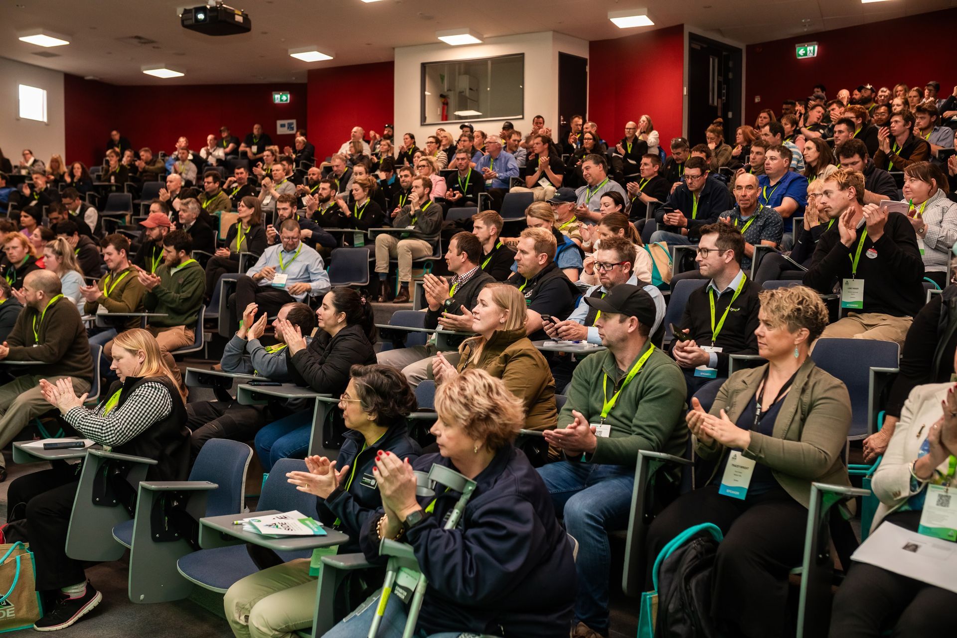Audience applauds in a large auditorium with red walls. People wear lanyards, some clap.