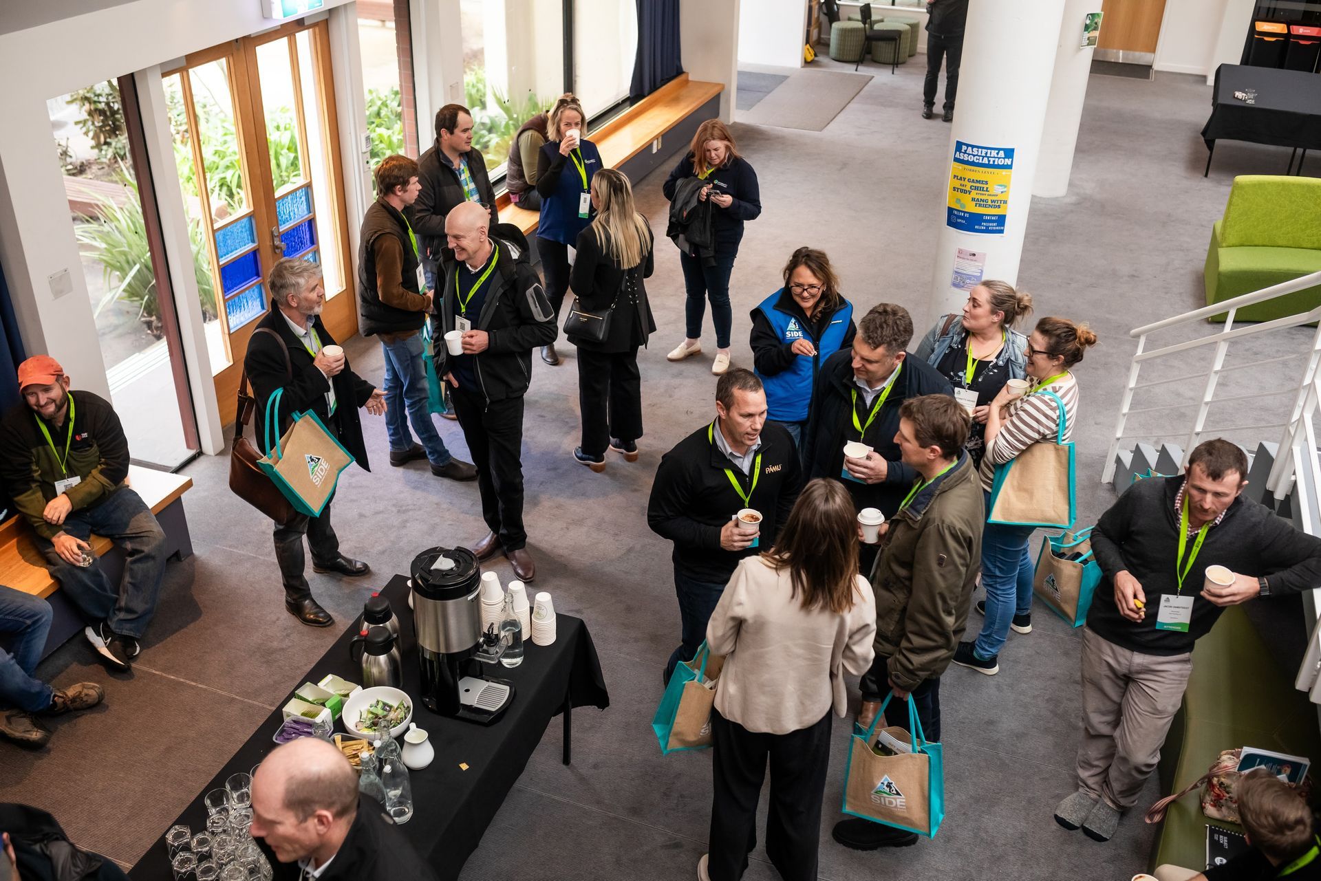 People at a networking event, gathered by a coffee station. Some are holding drinks, talking, and wearing lanyards.