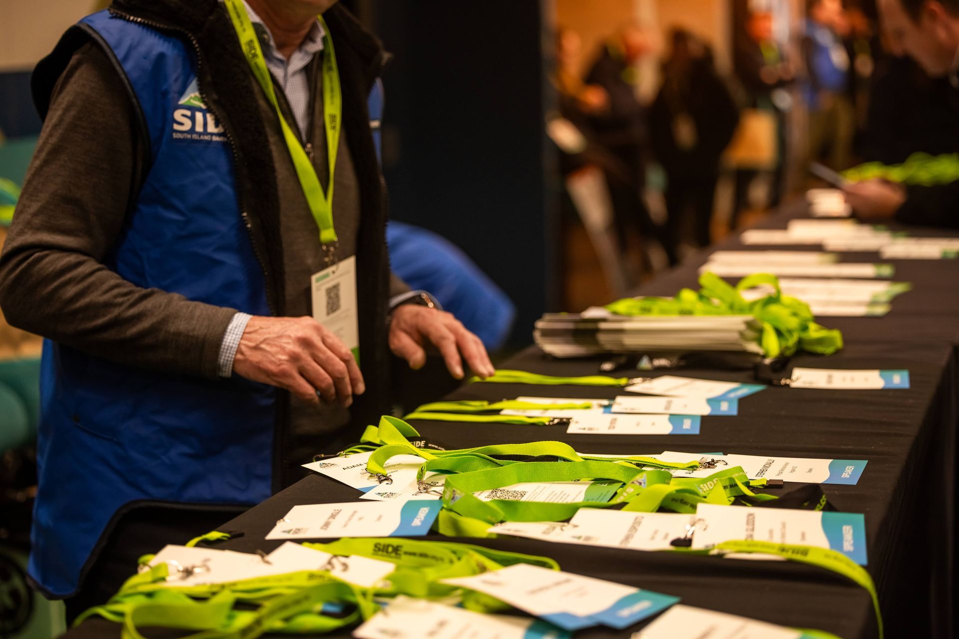 Person at a registration table, wearing a blue vest, handling name tags and lanyards.