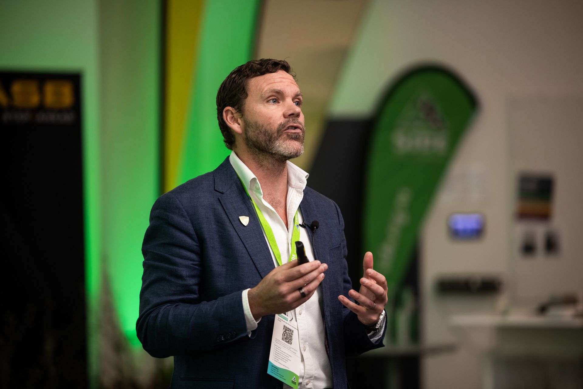 Man in blazer speaking at an event, gesturing with hands, green and yellow background.