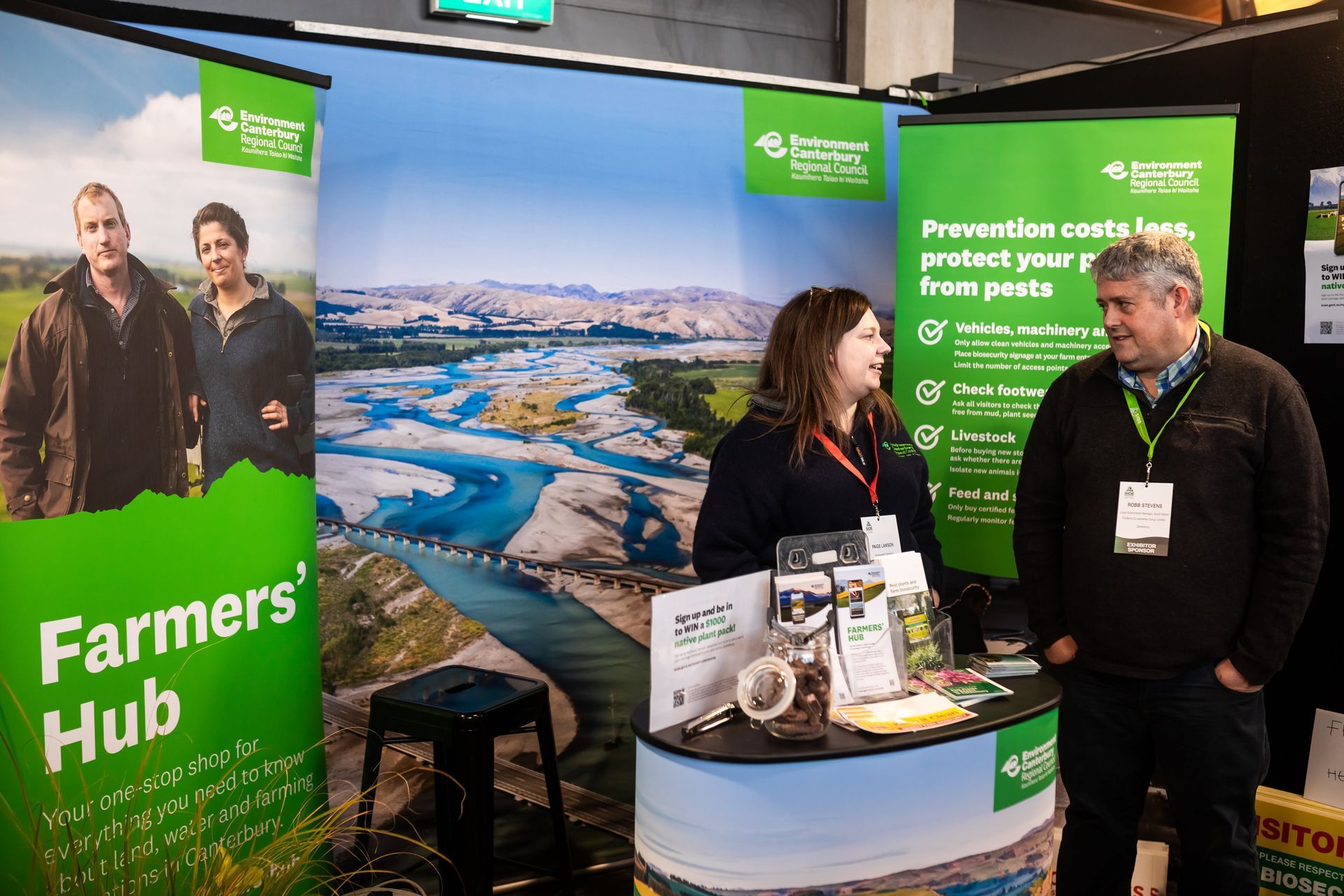 People at Farmers' Hub booth, discussing near promotional banners, with a river landscape backdrop.