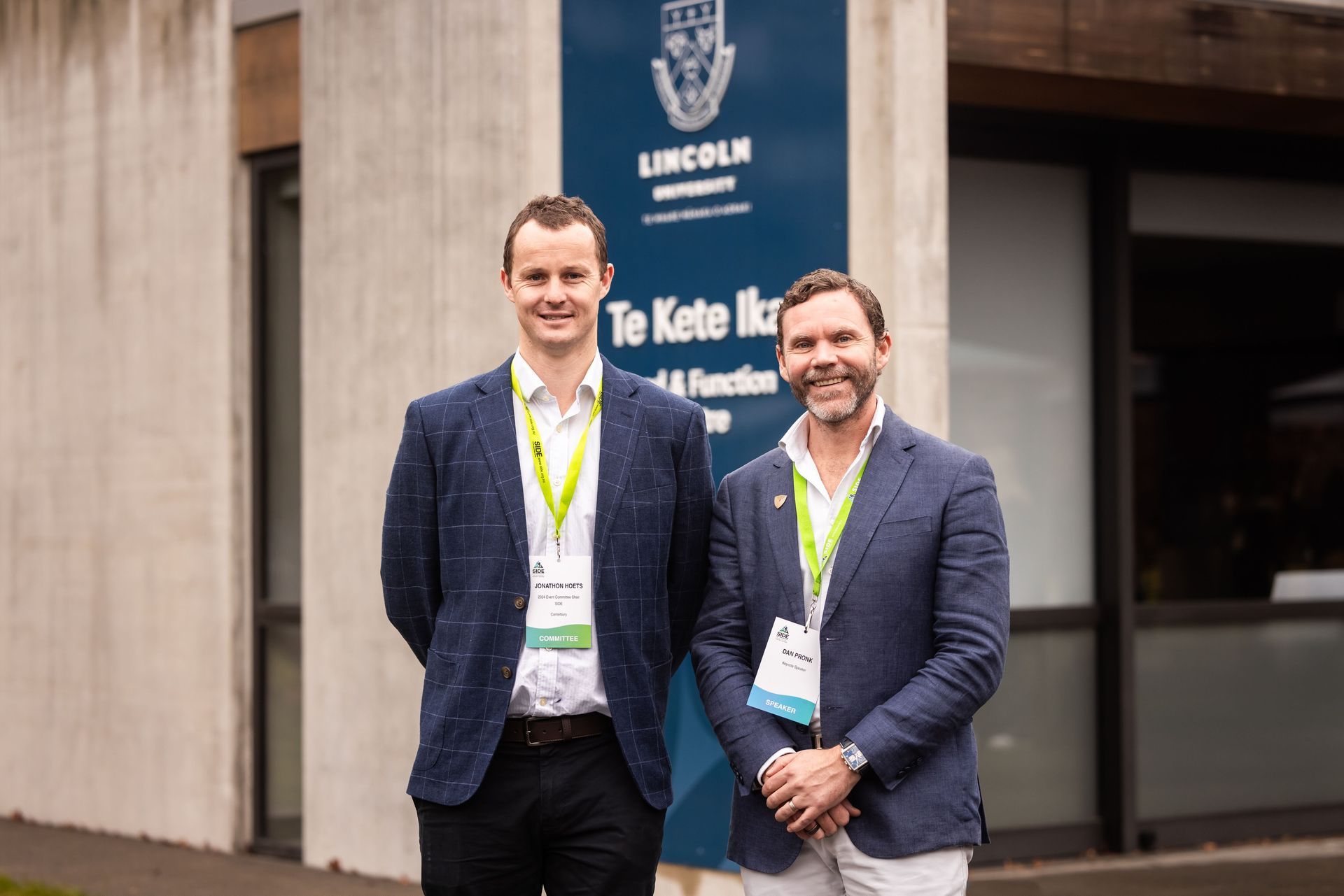 Two men in jackets stand in front of a building with a Lincoln University sign.
