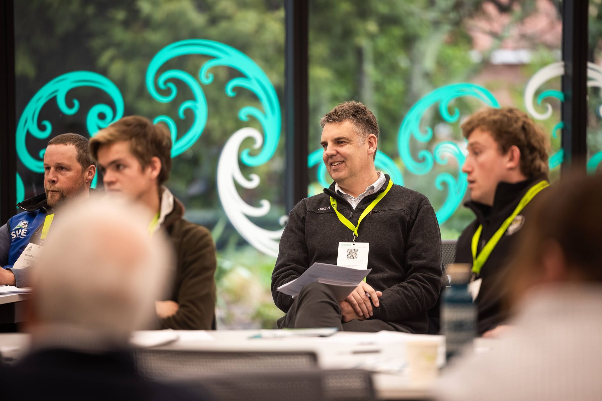 Group of people in a meeting, some wearing name tags. Turquoise swirls decorate the window behind them.