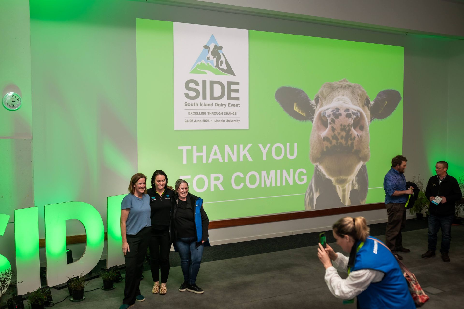 People posing in front of a green backdrop with the SIDE logo and a cow. A person takes a photo of them.
