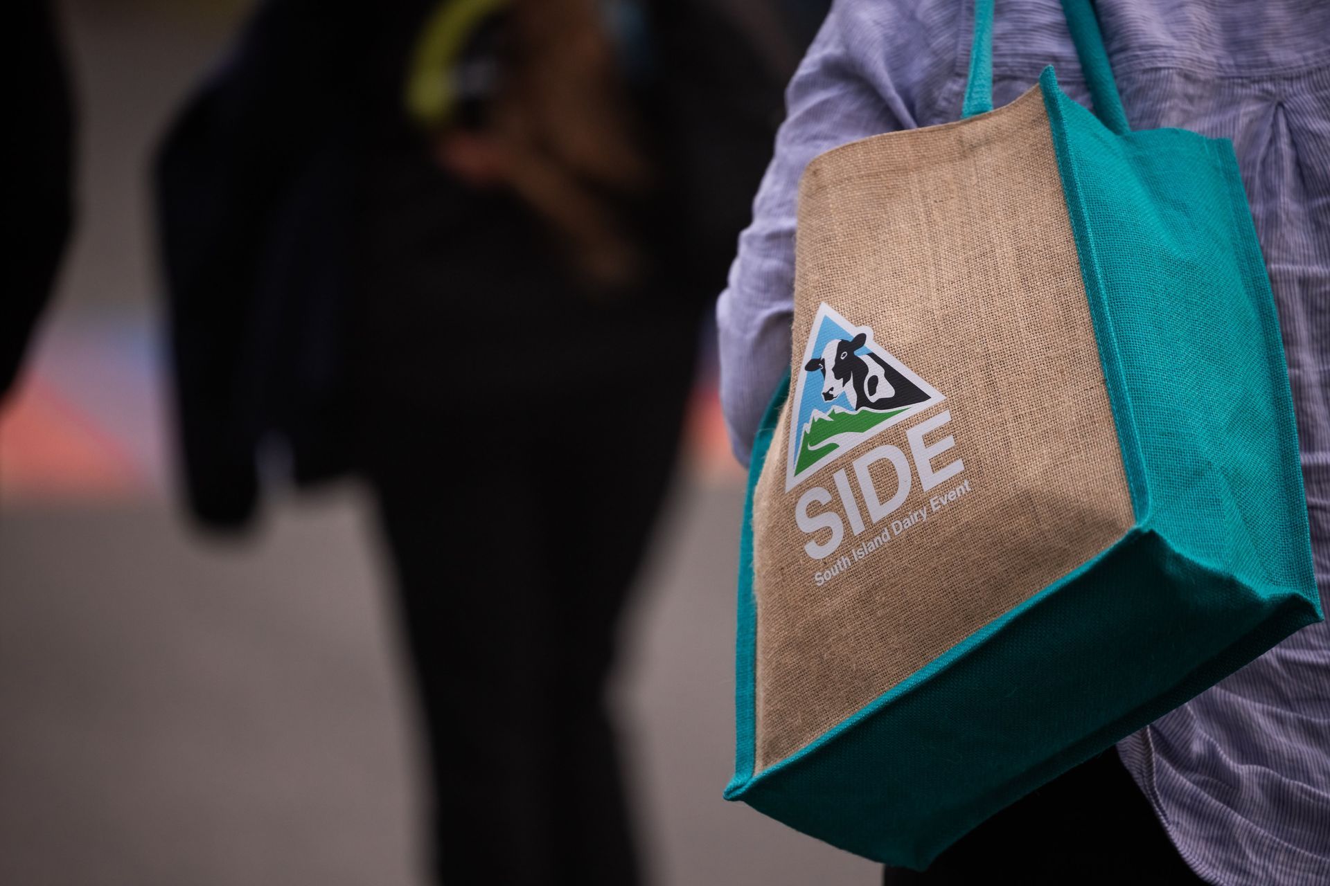 Person holding a tan tote bag with teal accents; the bag has a logo that says 